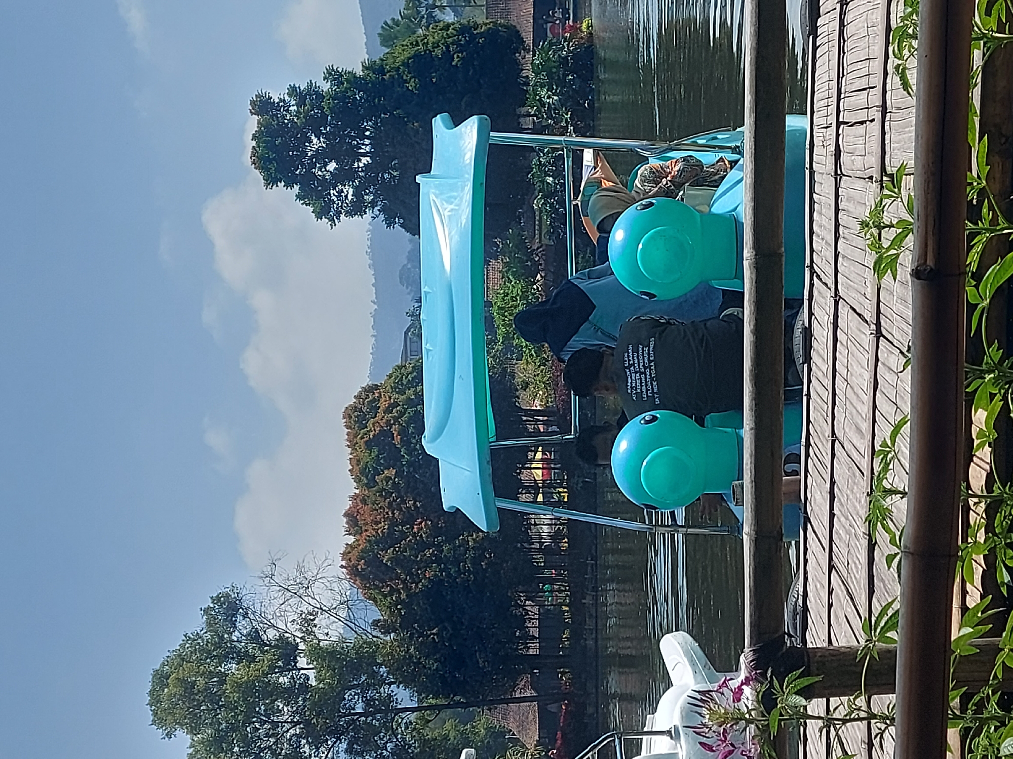 In Lembang, Indonesia, on a bright, sunny day, a group of people are enjoying a ride on a blue pedal boat shaped like a duck. The boat is on a calm body of water, with lush green trees and vegetation lining the shore. In the background, there are buildings with red tile roofs, characteristic of Indonesian architecture. The sky is a clear blue with a few scattered white clouds. The scene appears to be a recreational area, possibly a tourist attraction, where visitors can rent pedal boats and enjoy the natural scenery.