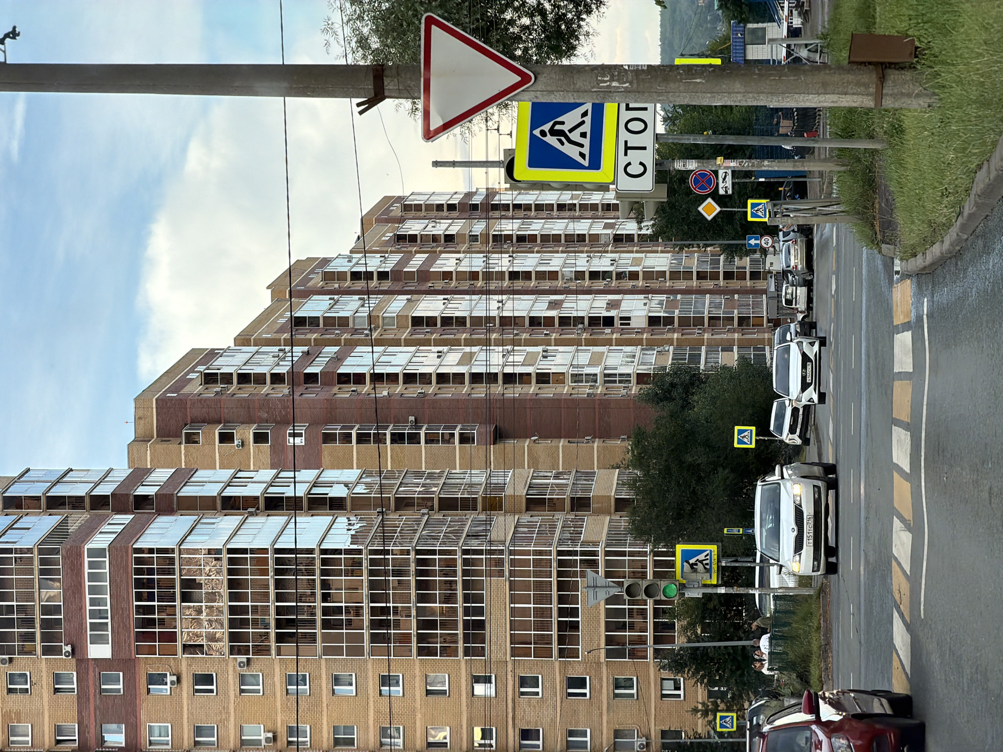 The image depicts a street scene in Kazan, Russia, characterized by modern, multi-story apartment buildings with brick and glass facades. Several cars are visible on the road, with one white car in the foreground driving towards a crosswalk. Traffic signals and various road signs, including a pedestrian crossing sign and a yield sign, are present. A concrete pole with multiple signs is prominently featured on the right side of the image, alongside green grass and a curb. The sky is partly cloudy.