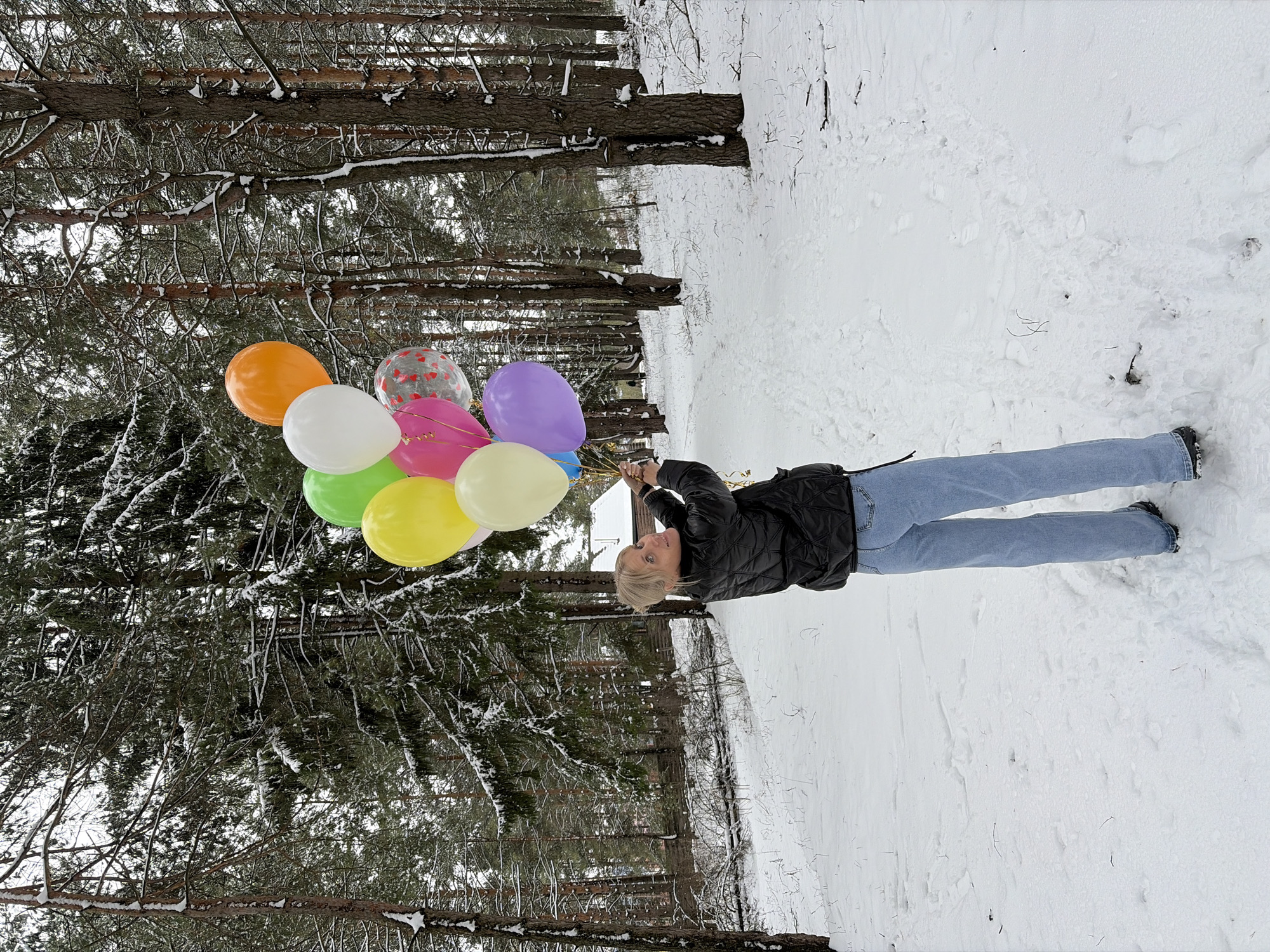 The image shows a woman holding a bunch of balloons in a snowy forest landscape. The woman is wearing a black jacket and blue jeans. The balloons are various colors including orange, white, clear with hearts, pink, purple, blue, green, and yellow. The trees are tall and snow-covered, creating a dense forest environment. The ground is covered in a thick layer of snow. The location is Прикамский, Russia, based on information provided about the image.