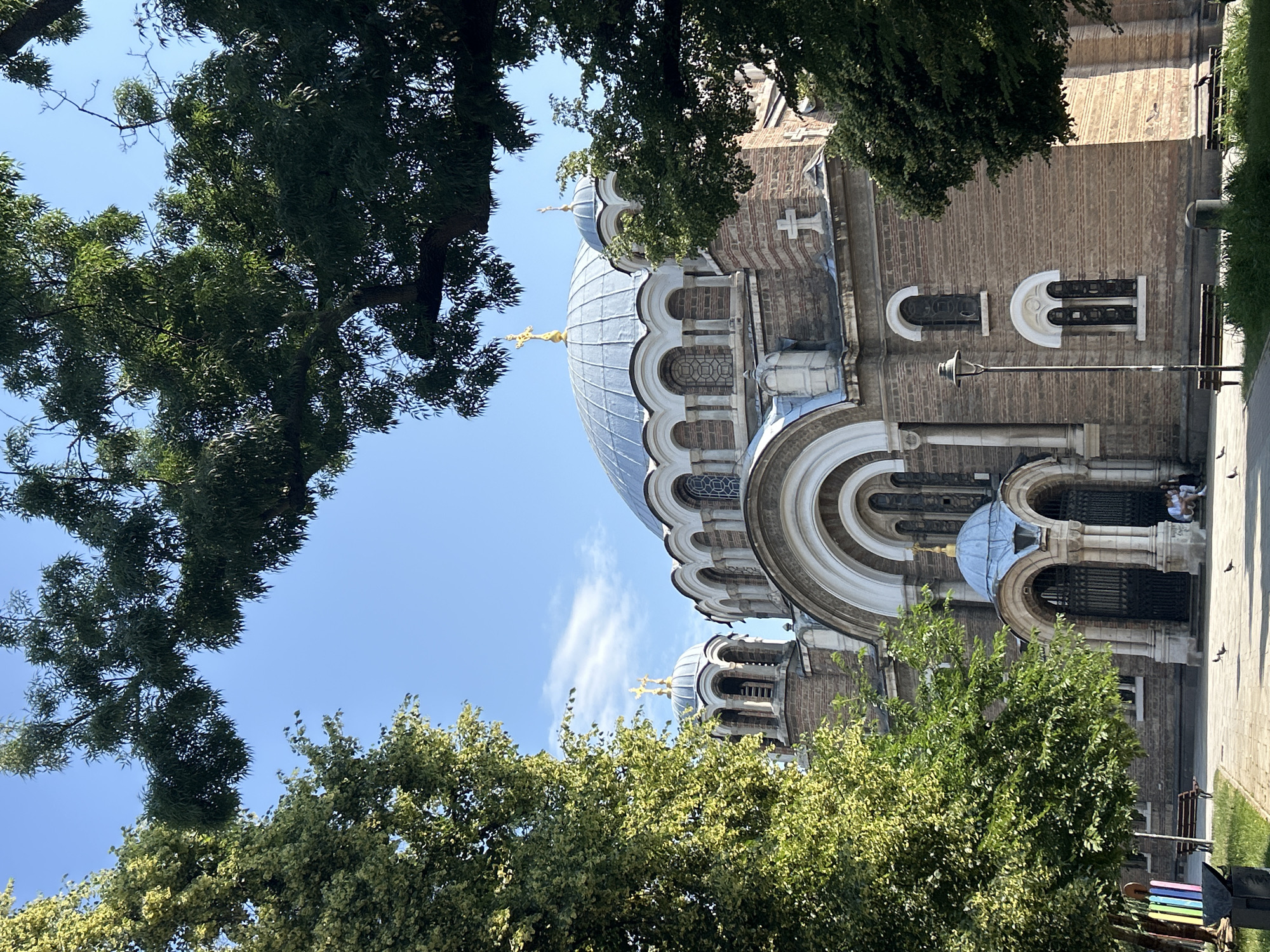 The image shows the Alexander Nevsky Cathedral in Sofia, Bulgaria. The cathedral is a large, Neo-Byzantine church with multiple domes, the largest of which is covered in gold. The building is constructed of brick and stone, with ornate architectural details. The scene is framed by lush green trees, suggesting a pleasant, sunny day. In the foreground, there is a paved area with benches and a lamppost. A few pigeons are walking on the ground, and two people are sitting on a bench near the entrance of the cathedral. The sky is clear and blue with a few wispy clouds.