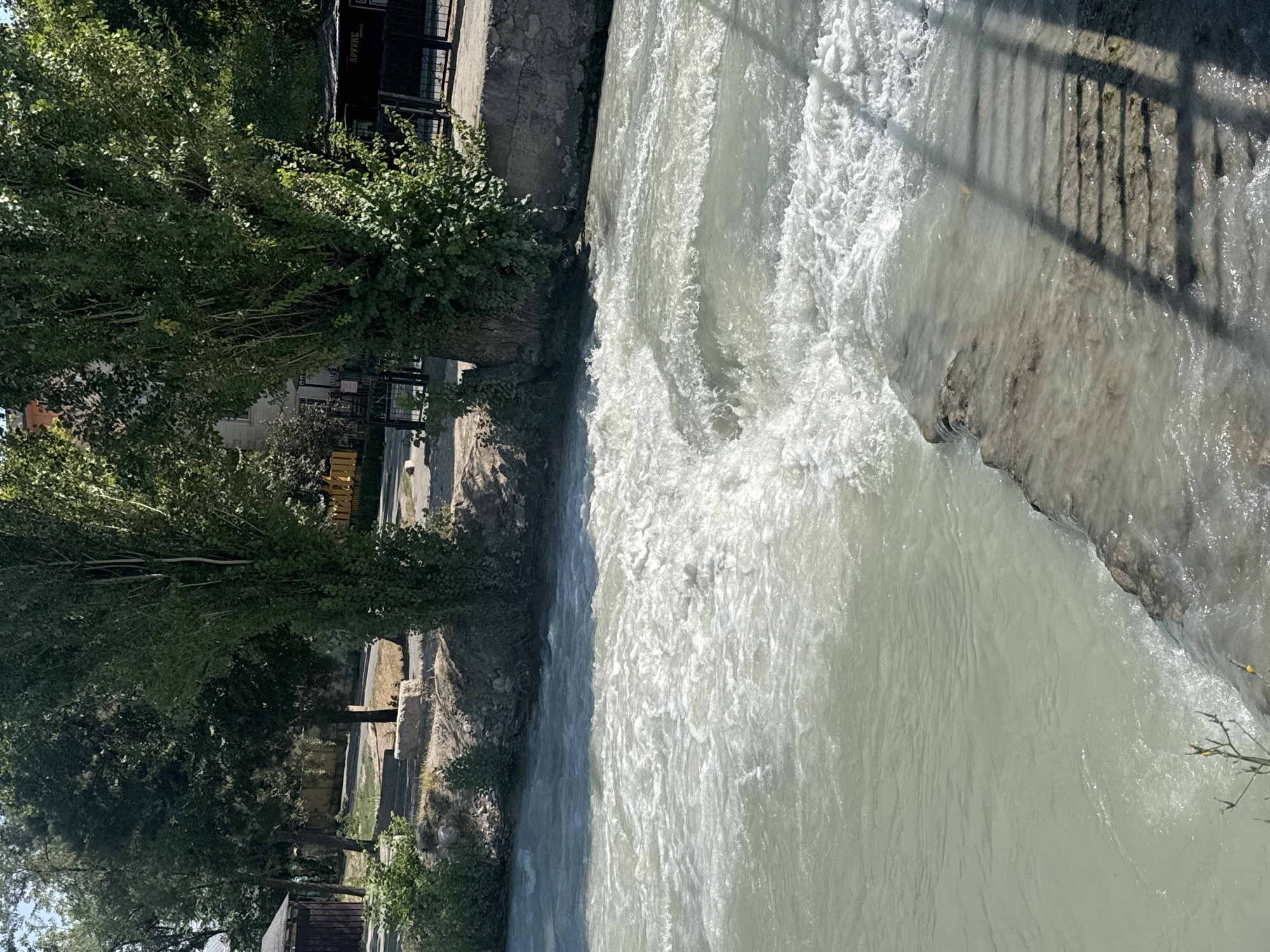 The image shows a view of a fast-flowing river in Bishkek, Kyrgyzstan. The river has white, turbulent water with small rapids, suggesting a strong current. On the left bank, there is a small wooden structure and lush green trees. The right bank is lined with a stone embankment, a black metal fence, and more trees. The ground on the banks appears to be paved or gravelly. The water surface reflects the bright daylight, and a shadow of a fence or railing falls across the lower right portion of the image, indicating the presence of a walkway or bridge. The overall scene depicts a natural setting within an urban environment, with the river being the central focus.