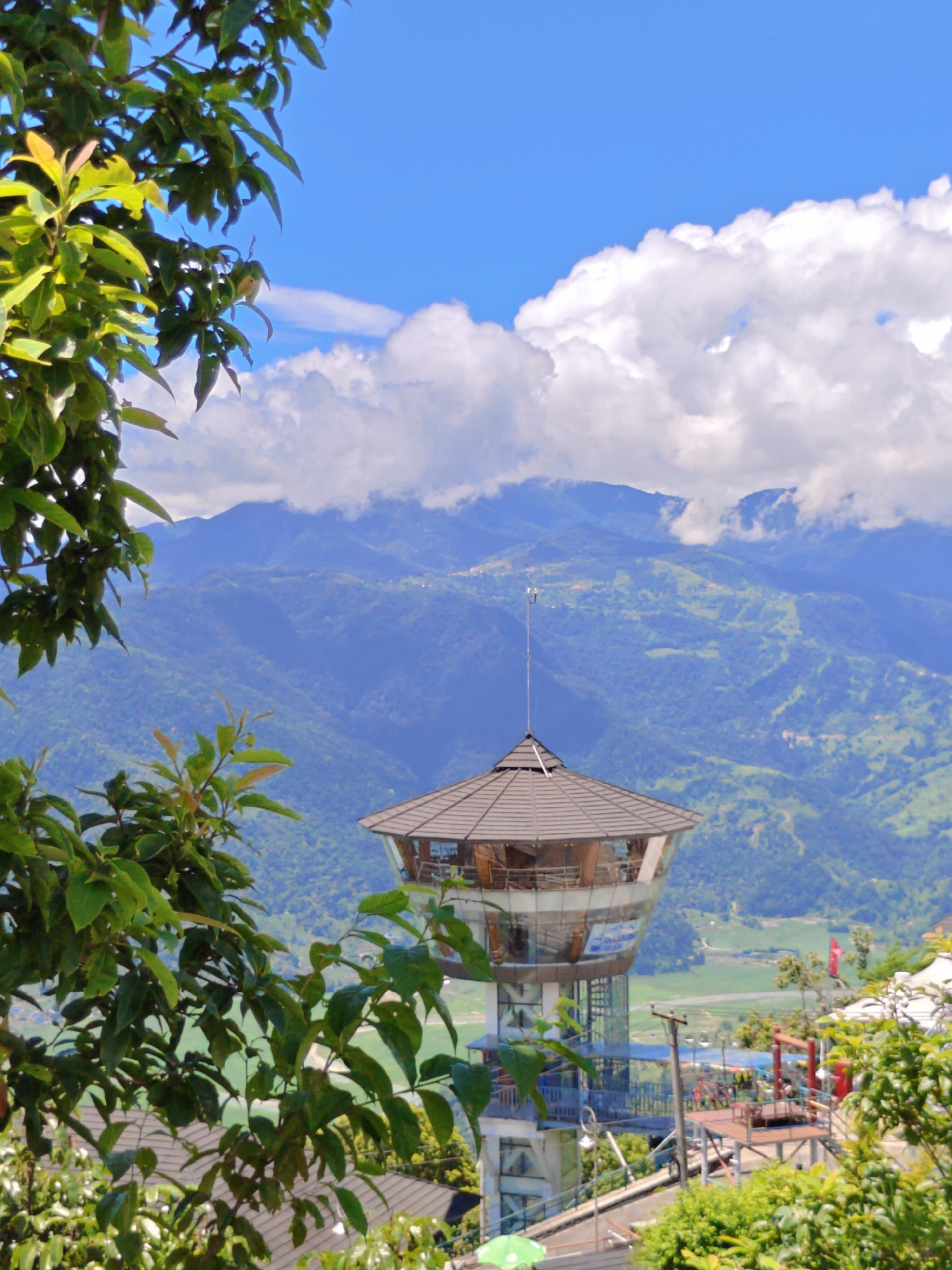 The image depicts a panoramic view from a vantage point in Pokhara, Nepal. In the foreground, lush green foliage frames the scene, with branches and leaves partially obscuring the view. Dominating the middle ground is a distinctive observation tower, featuring a glass-enclosed upper level with a hexagonal roof and a central antenna. The tower is supported by a multi-story structure with exposed beams and railings. Below and to the right of the tower, a complex of platforms and structures, possibly part of an adventure activity, is visible.
The background is characterized by rolling, verdant hills and mountains, some of which are shrouded in soft, white clouds. The landscape suggests a valley or basin, with patches of cultivated land and scattered buildings visible in the distance. The sky above is a vibrant blue, dotted with fluffy white clouds. The overall atmosphere is one of natural beauty and recreational development, with the observation tower serving as a focal point for appreciating the expansive scenery of Pokhara. No people are visible in the frame.