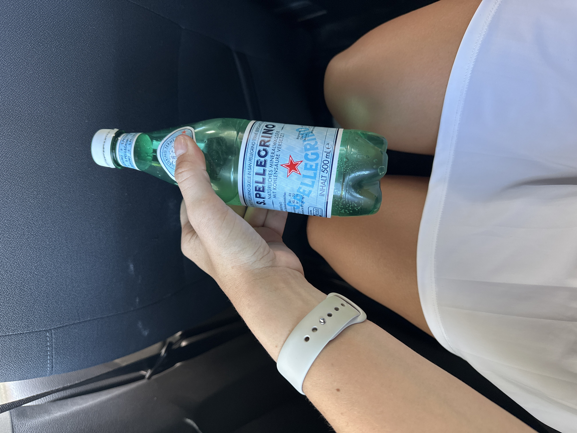 The image shows a person holding a bottle of S.Pellegrino sparkling mineral water. The bottle is green with a white cap and a label with the brand name and information in both Italian and German. The person is wearing a white watch band. The setting appears to be inside a car. The person is wearing white shorts or a skirt. The background includes a dark car seat. Dillfeld, Germany is not visible in the image.