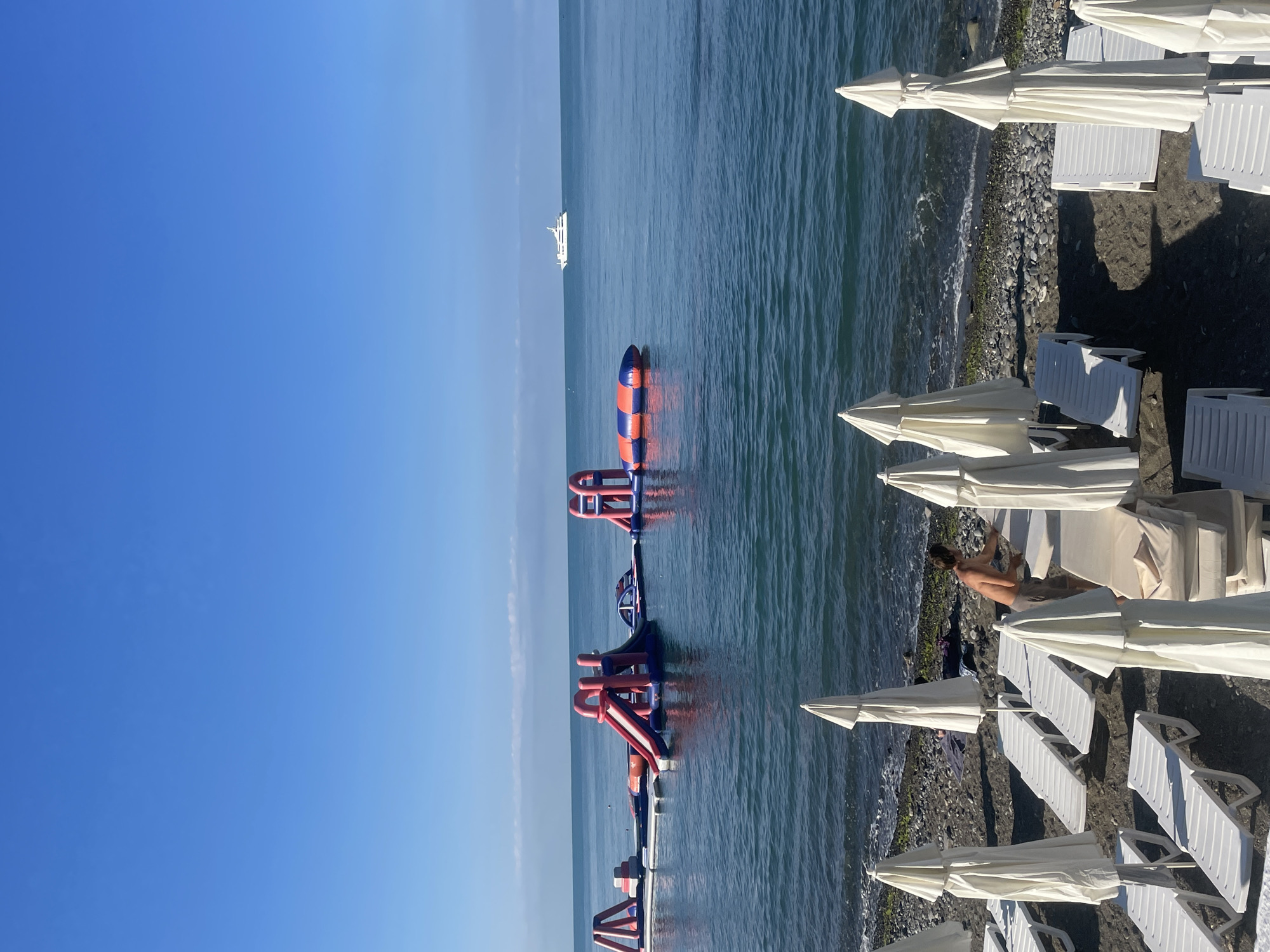 A sunny day at a Black Sea beach in Sochi, Russia, with an inflatable water park visible offshore and lounge chairs and umbrellas set up on the pebble beach. A lone man is seen arranging lounge chairs near the water. In the distance, a large white yacht sails on the calm blue sea under a clear sky.