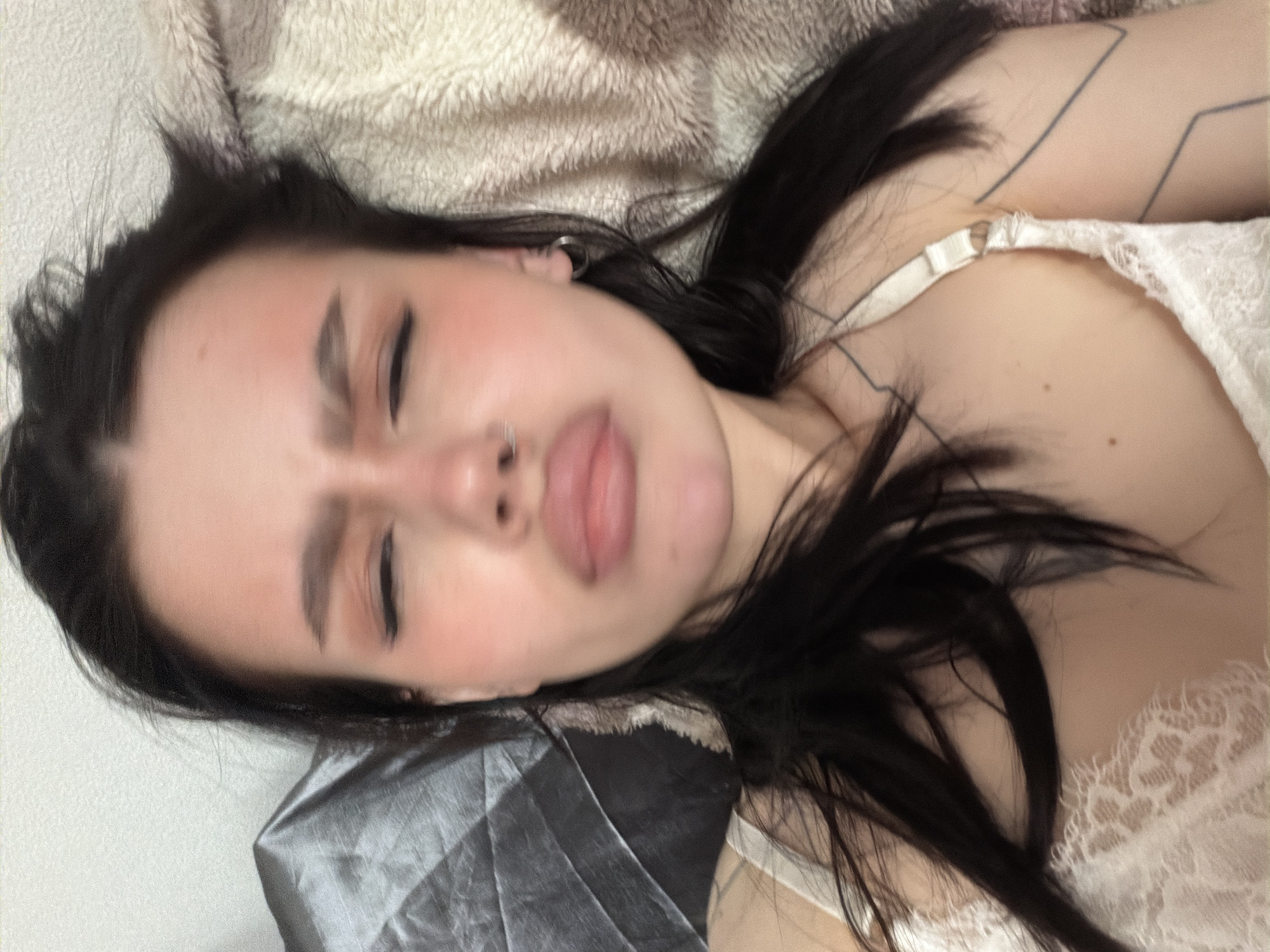 A woman with dark hair lies on a bed. She sticks her tongue out. The bedding appears to be a mix of light beige and silver-grey materials. She wears a white lace garment. A tattoo is visible on her arm. The location is Unknown, Unknown.