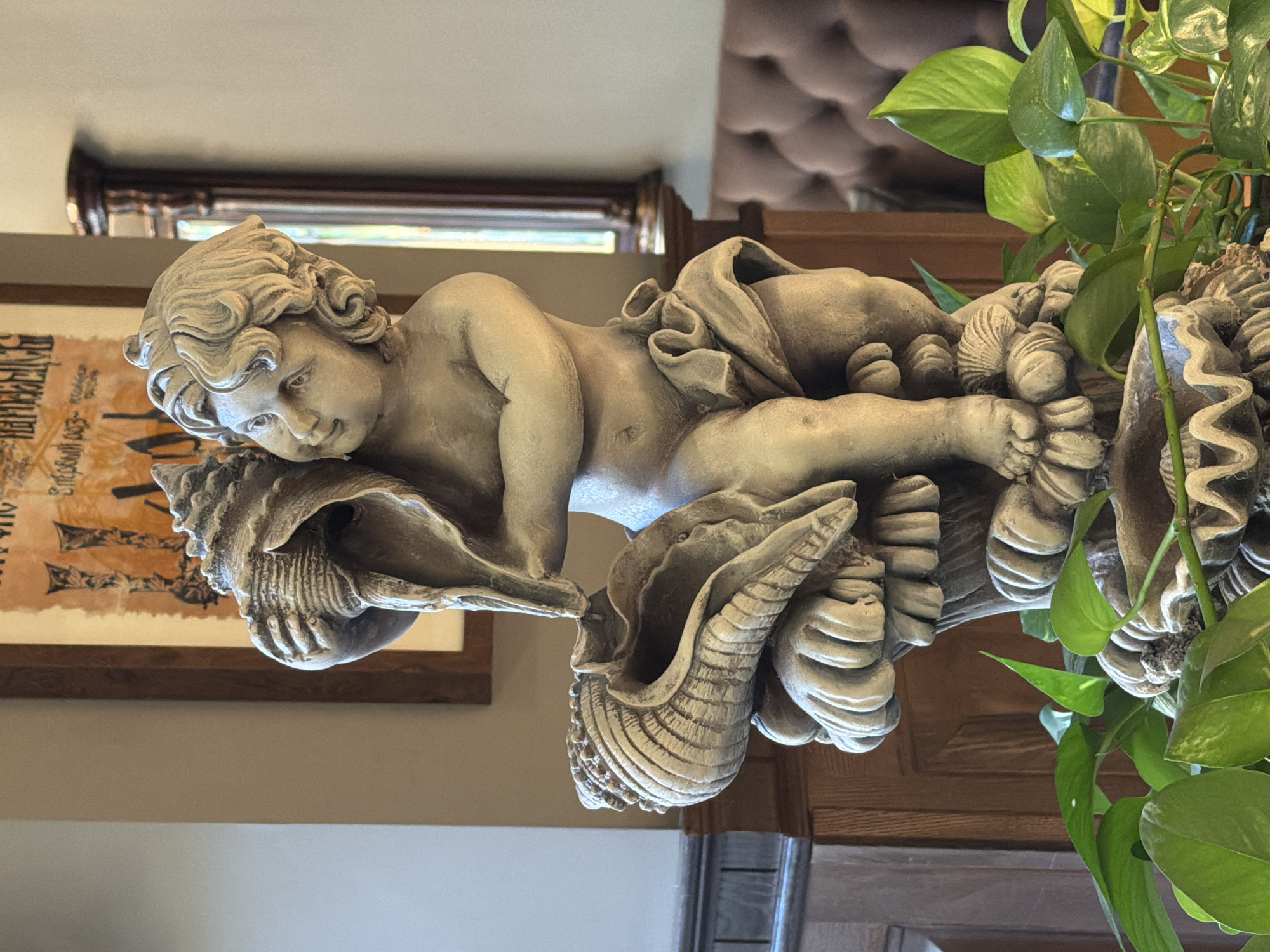 A stone statue of a cherub holding a conch shell is displayed in a room. The cherub is seated on a base adorned with more shells and wave-like patterns. In the foreground, green plants with broad leaves are visible. In the background, a framed artwork hangs on a beige wall, and a tufted brown couch can be seen, along with a dark wooden pillar and a tall, ornate mirror. The lighting suggests an indoor environment, possibly a home or a decorative establishment, in Rostov-on-Don, Russia.