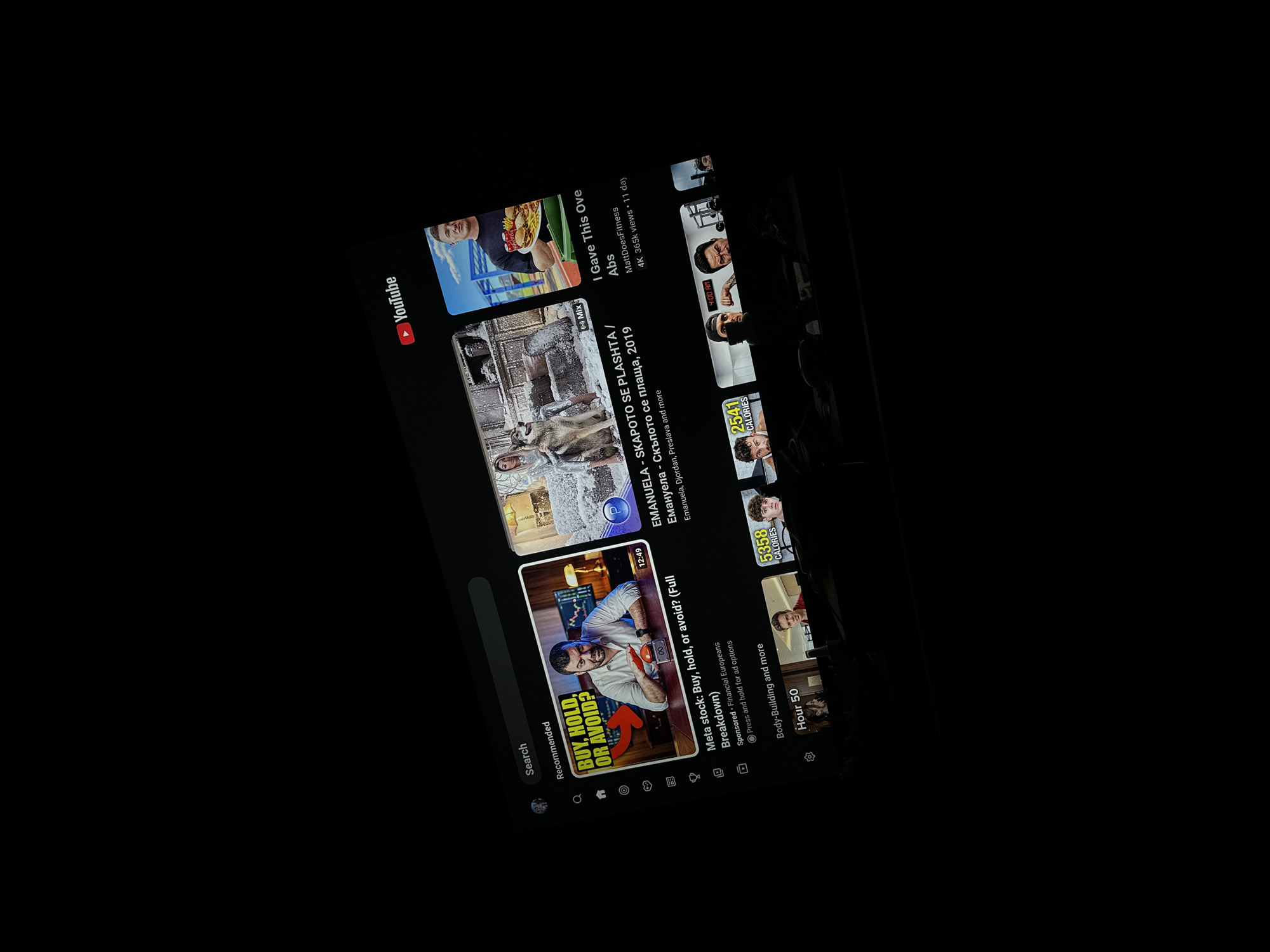 A mobile phone screen, displaying the YouTube application in dark mode, is captured at a diagonal angle in a dimly lit environment, likely indoors in Blagoevgrad, Bulgaria. The screen is the central focus, showcasing various video recommendations and parts of the YouTube interface.
Prominently featured on the left is a video thumbnail with a man in a white shirt and text "BUY, HOLD, OR AVOID?". The accompanying title reads "Meta stock: Buy, hold, or avoid? (Full Breakdown)," marked as "Sponsored • Financial Europeans" and indicating a duration of "12:49."
Above this, another video thumbnail shows a woman and a large dog or wolf in a snowy landscape, with the title "EMANUELA - SKAPOTO SE PLASHTA / Емануела - Скъпото се плаща, 2019" by "Emanuela, Djordan, Preslava and more." To the top right, a video from "MattDoesFitness" is titled "I Gave This Ove Abs," showing "4K 365k views • 11 day." Below this, smaller thumbnails feature fitness content, including text like "5358 CALORIES," "2541 CALORIES," "Body-Building and more," and "Hour 50," with one also displaying "4:00 AM."
The standard YouTube logo is visible on the upper left, along with a "Search" bar and "Recommended" section label. Navigation icons for Home, Shorts, Create, Subscriptions, and Library are arranged at the bottom of the screen.