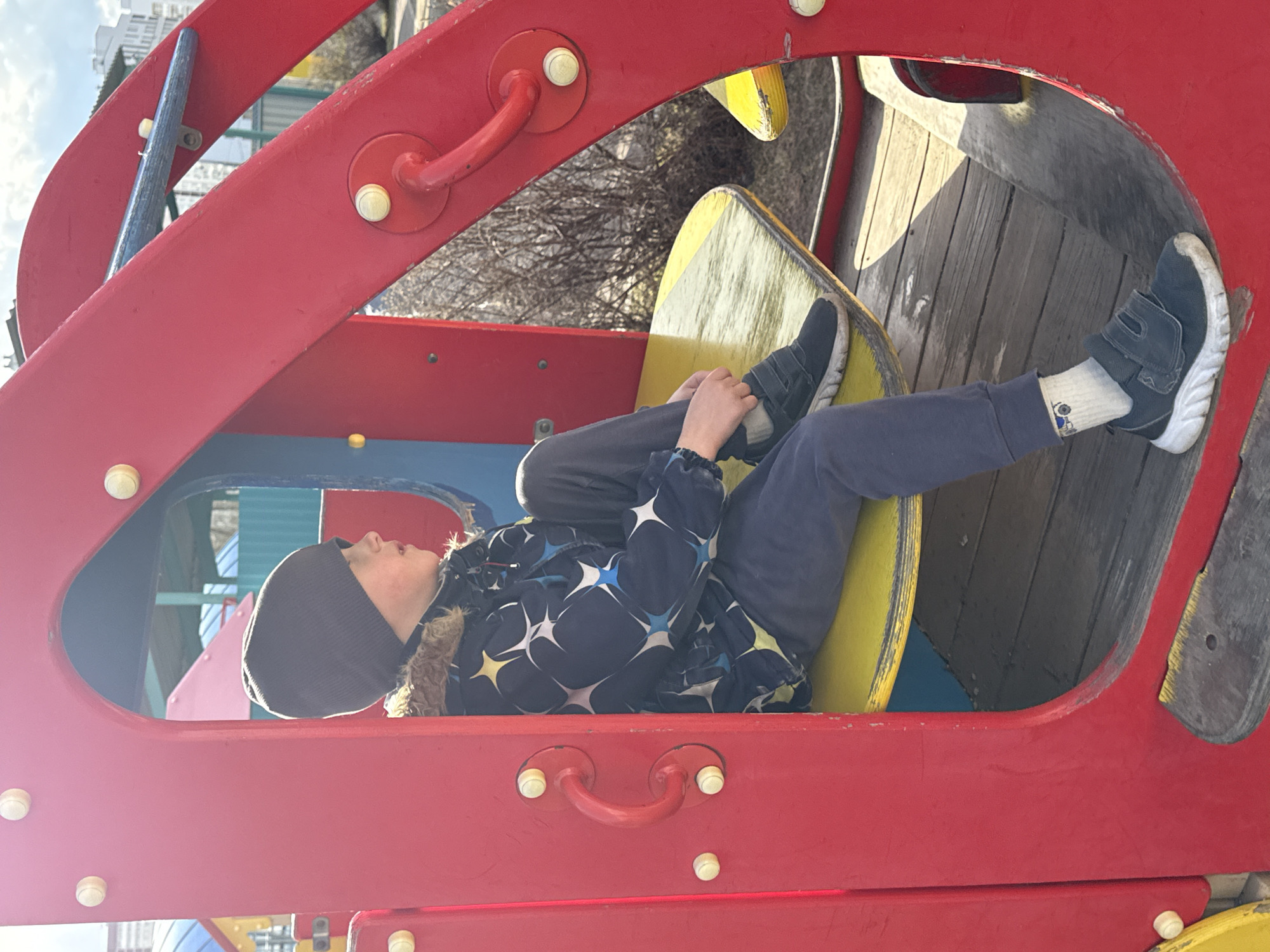 A young child is depicted lying on their back within a vibrant red and yellow playground structure on a bright, sunny day in Yekaterinburg, Russia. The child is wearing a dark grey beanie, a dark blue winter jacket patterned with white and yellow stars and a fur-lined hood, dark blue trousers, and dark athletic shoes with white soles and velcro straps. White socks, with a small patterned detail that resembles a robot or vehicle, are visible above their shoes. The child appears relaxed, with eyes closed or looking upwards, possibly resting or gazing at the sky.
The playground equipment is characterized by its bold colors: a prominent red frame, likely made of metal or painted wood, forms the arched supports and enclosing structure. Inside, bright yellow wooden or plastic platforms serve as seating or surfaces for play, with additional wooden planking forming part of the structure's base. White or light yellow circular fasteners and red handles are integrated into the red framework.
The scene is illuminated by strong, direct sunlight, casting distinct shadows and indicating clear weather. In the blurred background, bare tree branches and hints of urban buildings are visible against a bright sky, confirming an outdoor, urban park setting. No visible text is present in the image. The photograph is taken from a side angle, presenting the child horizontally across the frame.