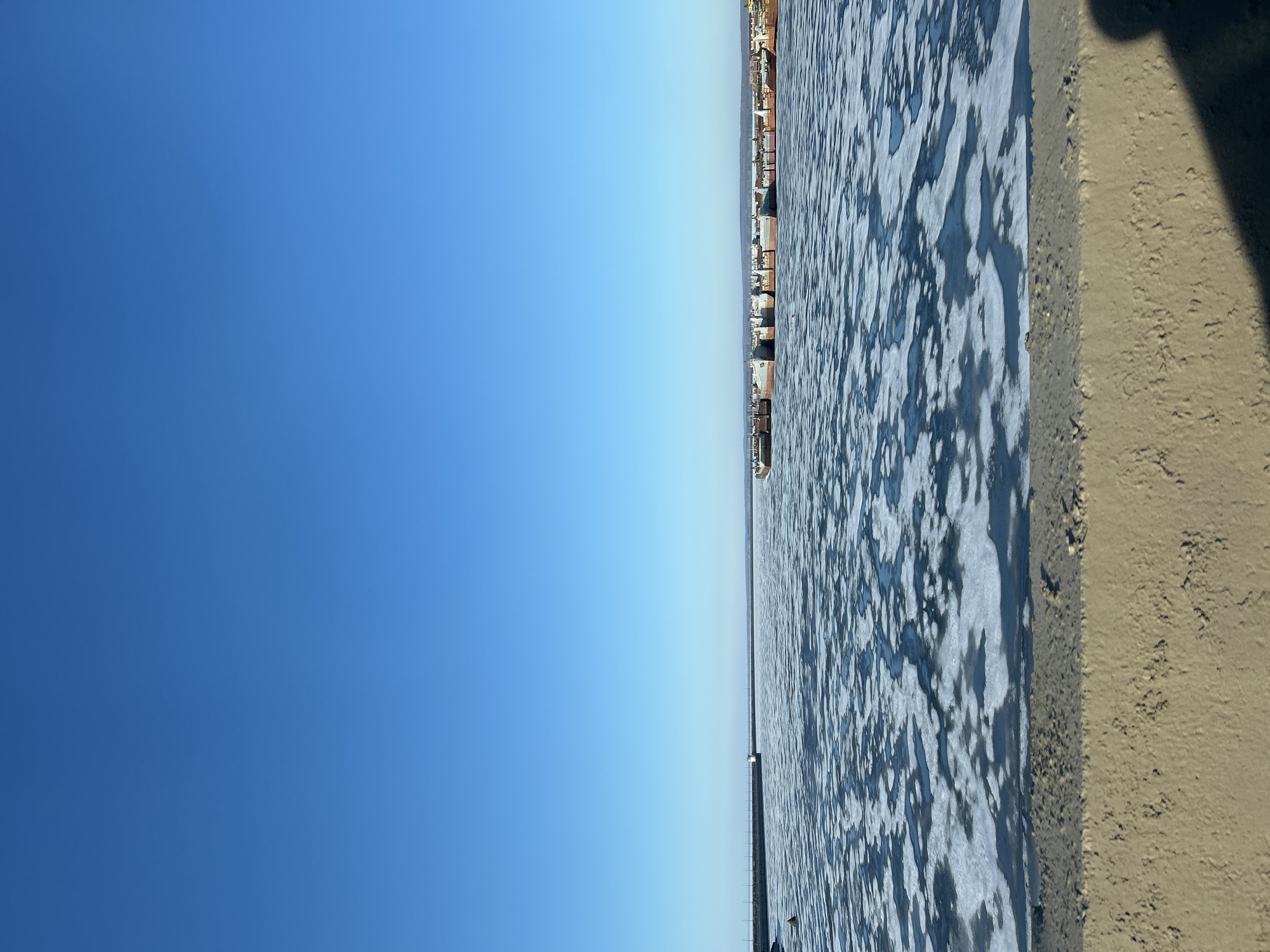 The image presents a horizontal view with the sky dominating the left side, transitioning into a body of water on the right, possibly the Kama River in Chaikovsky, Russia. The water shows signs of ice or foam patches. A line of buildings is visible on the horizon across the water. A section of embankment or riverbank is seen in the lower right corner of the image. The sky is clear and blue. The light suggests it's daytime. No people are directly visible in the image.