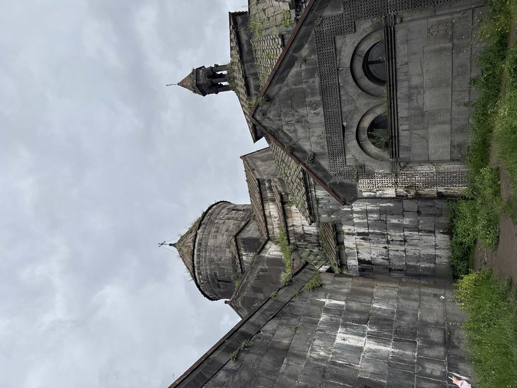 This is a photograph of the Haghpat Monastery in Haghpat, Armenia. The image focuses on the stone architecture of the monastery, with its large stone blocks, domed roofs, and arched windows. Vegetation, including grass and small plants, is growing on and around the structures. The sky is overcast with clouds, casting a diffused light on the scene. The perspective is from a low angle, looking up at the monastery. No people are prominently featured in the shot, though a small hand with a pink and white striped sleeve is visible near the bottom left of the frame, seemingly pointing. The overall impression is one of ancient, weathered stone against a gray sky.