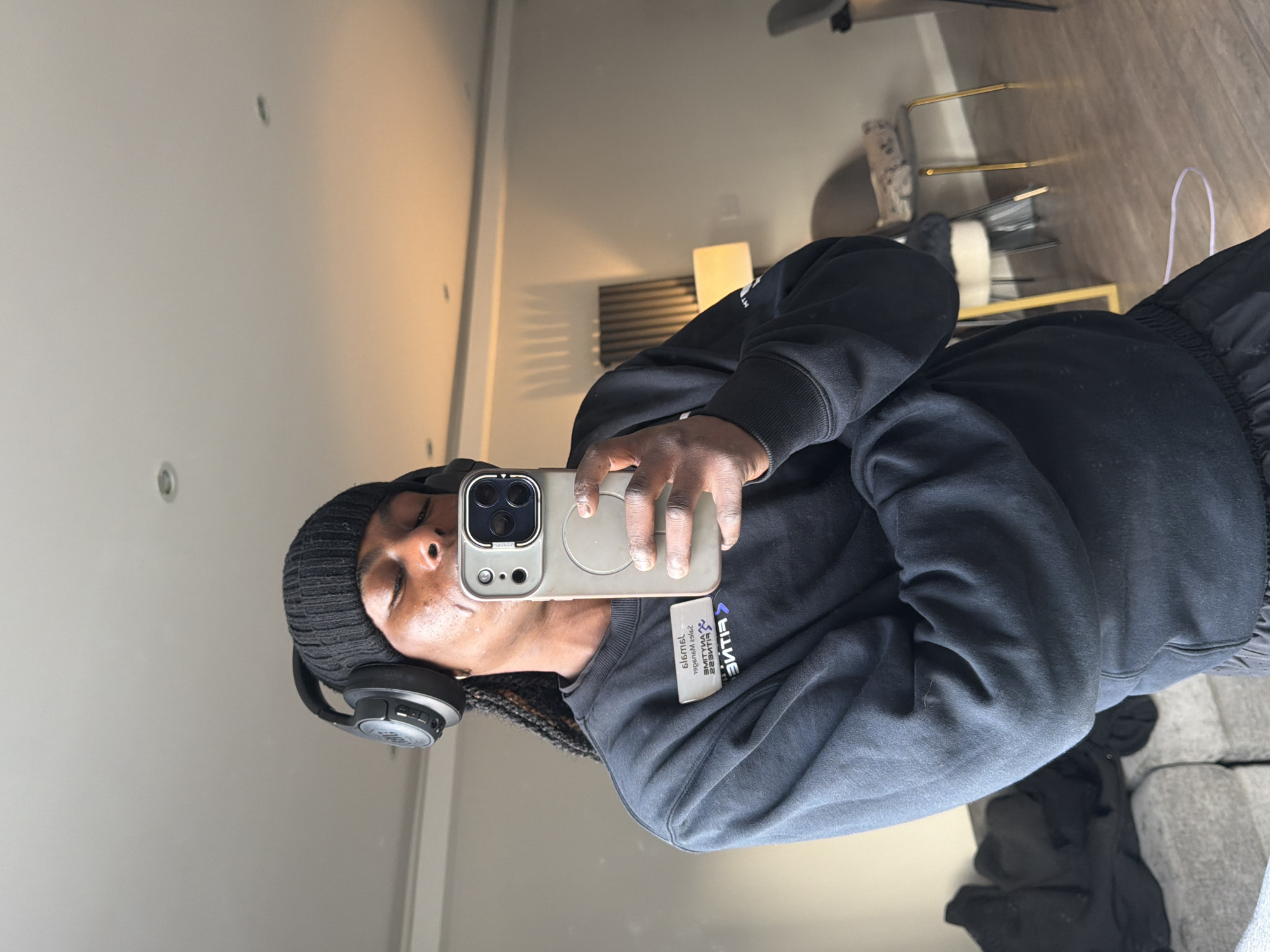 A person is taking a selfie in a room with neutral-colored walls and a light-colored ceiling. The person is wearing a black beanie, headphones, and a black sweatshirt with a "Lifetime Fitness" name tag attached. They are holding a smartphone with a light gray case. A chair with a white seat and gold legs, along with a light fixture, are partially visible in the background. The image does not contain enough information to determine the location in London.