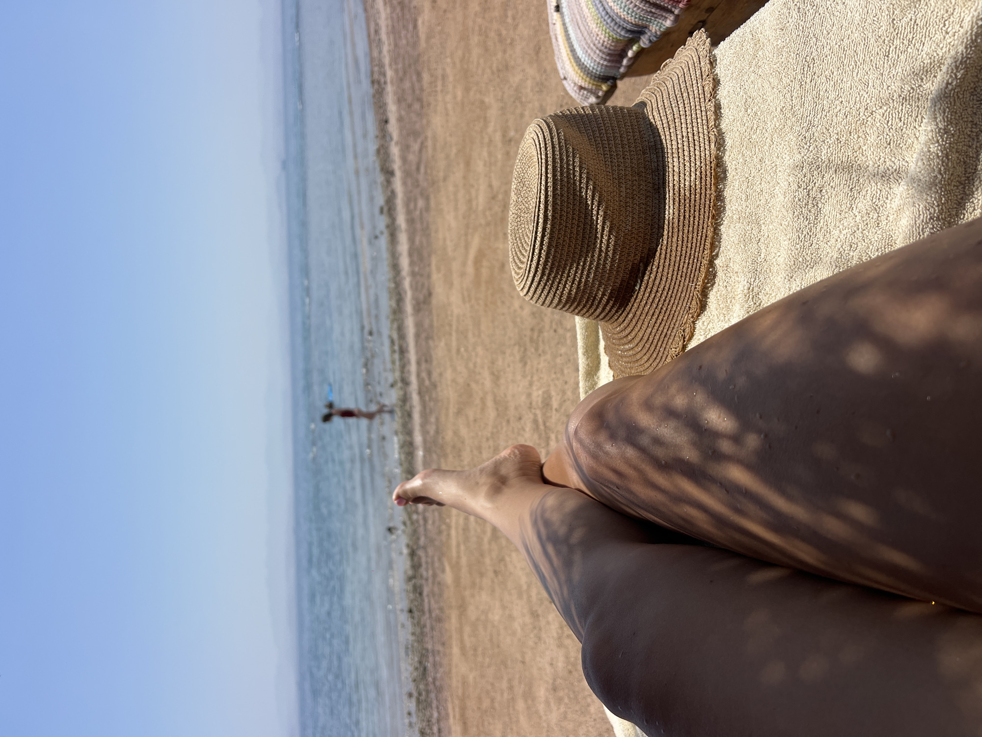 The image shows a first-person perspective of someone relaxing on a beach lounger on a sunny day in Nueiba, Egypt. In the foreground, the legs of the person are visible, along with a straw hat placed on a white towel. In the middle ground, the sandy beach extends to the edge of the clear blue sea. In the background, a person is wading in the shallow water, and the distant hazy outline of mountains can be seen against a clear blue sky. The scene depicts a peaceful beach vacation setting.