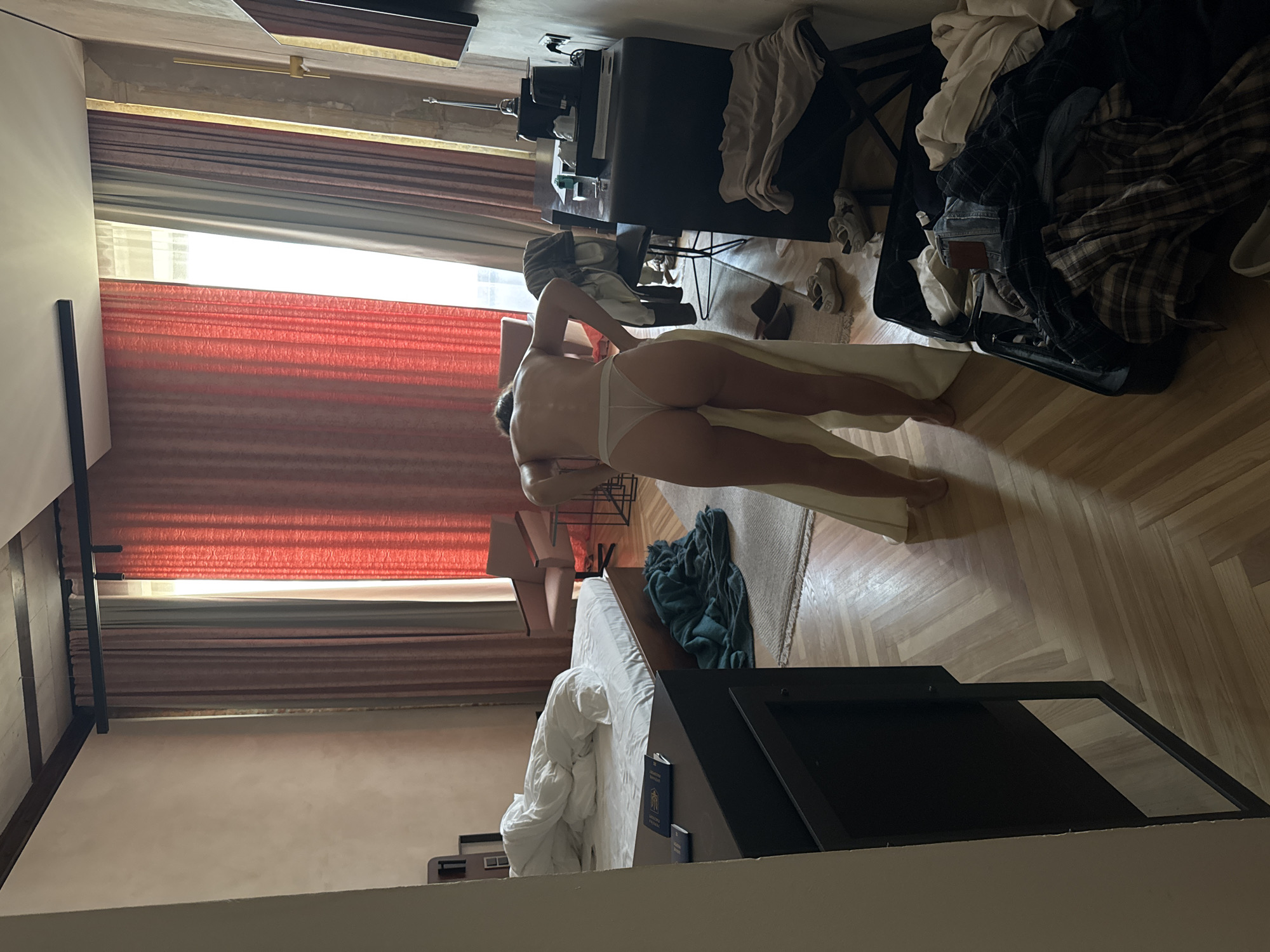 The image depicts the interior of a hotel room in an unknown city and location. In the foreground, a person, identified as a woman, stands facing away from the viewer, partially dressed in white thong-style underwear. Her back and buttocks are visible, and she holds a light-colored garment, appearing to be in the process of dressing or undressing.
The room features a wooden floor with a herringbone pattern and a light-colored rug in the center. On the left side, an unmade bed with white sheets and a duvet occupies a significant portion of the space. Adjacent to the bed, a dark nightstand holds two passports, one blue with a visible gold emblem and the other darker in color. A dark teal-colored blanket or throw is casually draped on the floor near the bed.
Further into the room, on the right, an open suitcase filled with various items of clothing, including plaid shirts and jeans, rests on the floor. Several pairs of shoes, including white sneakers and dark slides, are scattered around the suitcase and near the woman. A dark desk or console is positioned on the right, topped with what appears to be a coffee machine, cups, and other small items. A dark chair is pulled up to this desk, with a garment draped over its back.
The background is dominated by large windows covered with floor-to-ceiling curtains in shades of red/orange and brown/grey, with sheer white curtains behind them, allowing abundant natural light into the room. A light-colored armchair is visible near the center of the background. The ceiling features exposed dark wooden beams on the left side. The walls are light-colored and appear to have a textured finish.