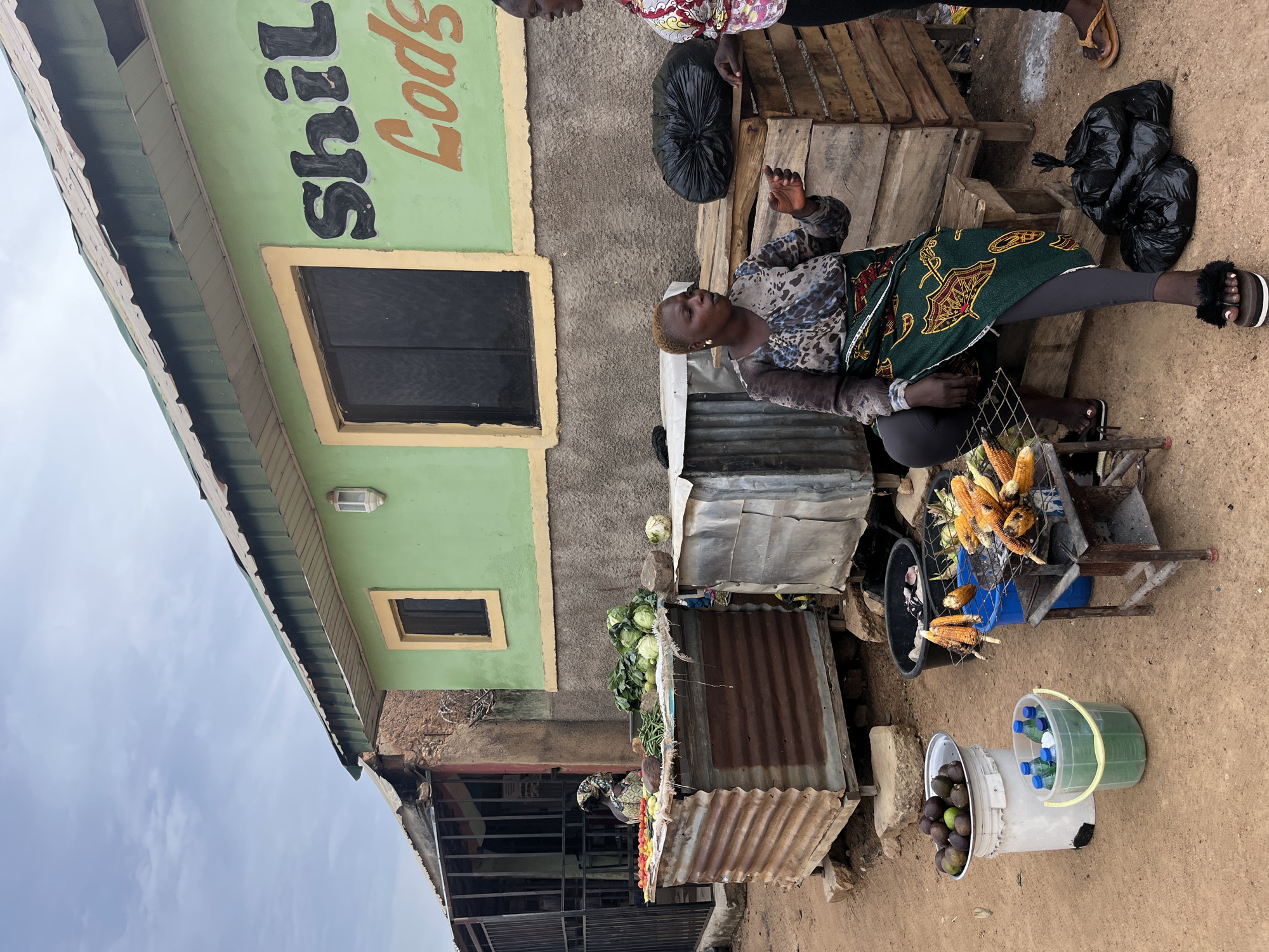 A woman is grilling corn at a roadside stand in Jos, Nigeria. She is sitting on a stool in front of a green building with "Shiloh Foods" painted on it in large black and orange letters. The building has a corrugated metal roof and a yellow trim around a dark window. To the left of the building is a corrugated metal stall displaying various vegetables, including cabbage, tomatoes, and green beans. Another woman, wearing a colorful patterned shirt, stands to the right of the grilling woman, engaged in conversation. In the foreground, several ears of corn are roasting on a metal grill over hot coals. Buckets containing avocados and bottled drinks are also visible. The ground is dusty earth, and the overall scene depicts a typical street market in Jos.
