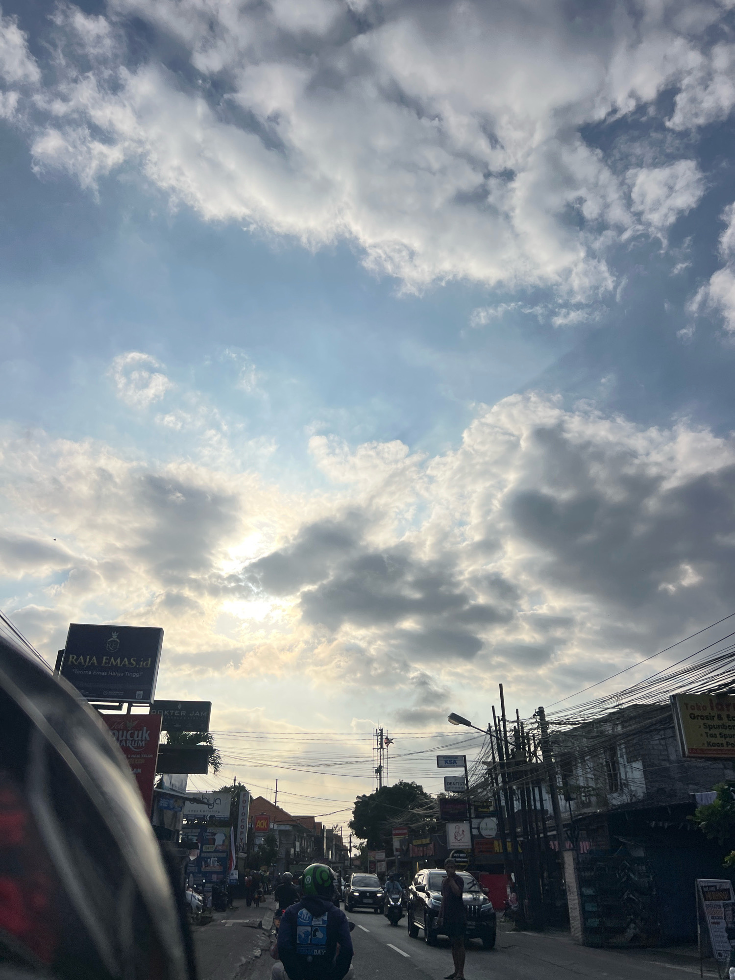 The image captures a street scene in Denpasar, Indonesia, under a dramatic sky filled with a mix of bright sunlight and dark, textured clouds. The sun is partially obscured by clouds, casting a warm, diffused light over the city. The foreground is dominated by the perspective of a vehicle, likely a motorcycle, with a portion of its dark, reflective helmet visible on the left.
The street is lined with buildings and various signs, suggesting a commercial area. Prominent signs include "RAJA EMAS.id" with a tagline in Indonesian, "DOKTER JAM," "Toko laris Grosir & Ecer," and others that are partially legible. Power lines are strung across the street, creating a dense network above the buildings.
Traffic is present, with cars and motorcycles on the road. A few individuals are visible, including a person in a green helmet on a motorcycle, another person standing near a dark SUV on the right side of the road, and others walking or riding in the distance. The architecture of the buildings appears to be typical of Southeast Asian urban environments, with some appearing older and others more modern. The overall atmosphere is that of a bustling urban street during the day, with the sky adding a sense of dynamic weather.