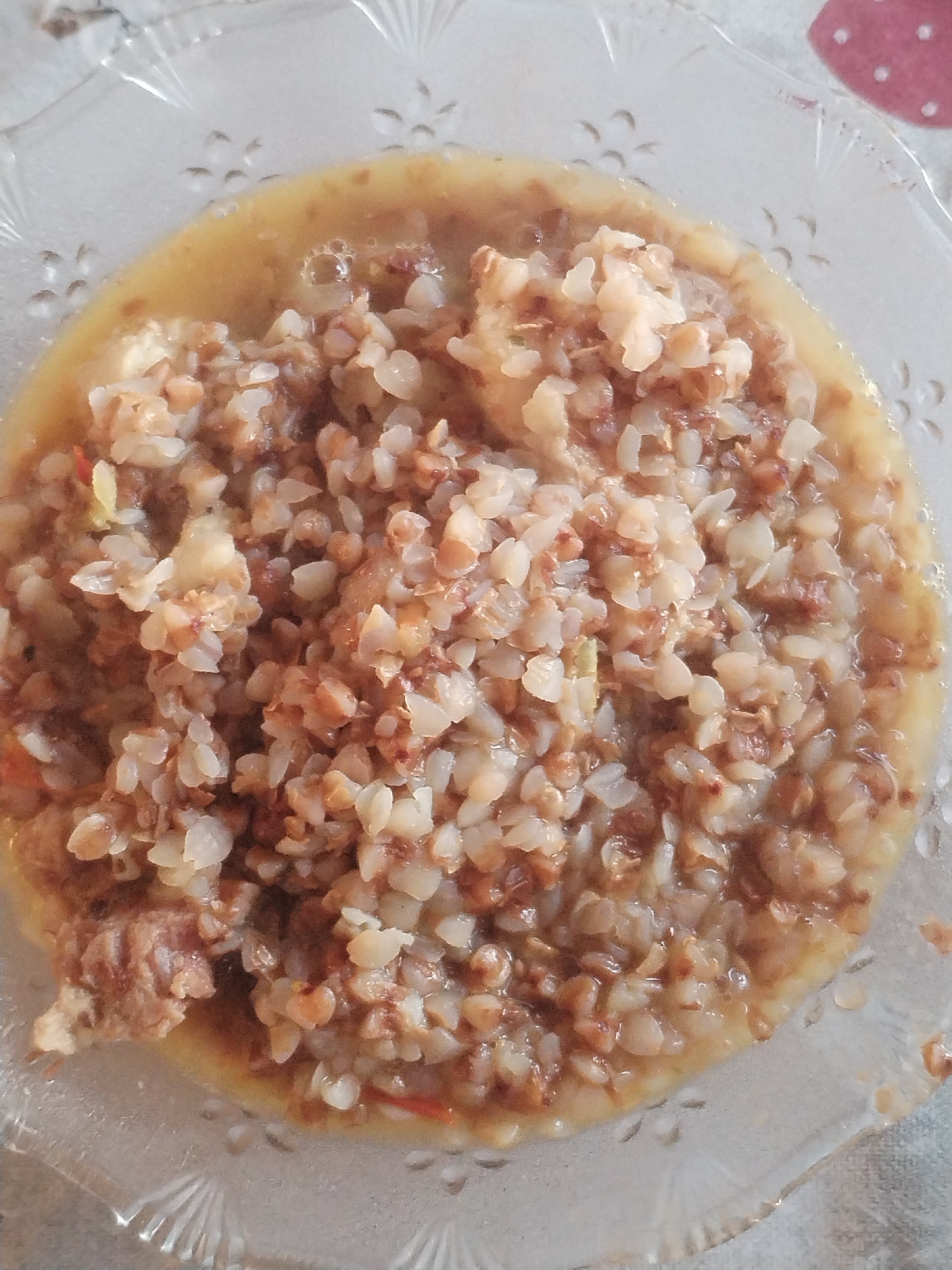 A close-up, overhead shot shows a bowl of buckwheat with meat. The buckwheat is a light brown color, with some grains appearing lighter and some darker. Mixed in with the buckwheat are pieces of meat, some of which are shredded. The dish is in a clear glass bowl with a fluted edge. The bowl is placed on a textured surface that appears to be a tablecloth. The background is out of focus but suggests a domestic setting. The image was taken in Moara Domnească, Romania.