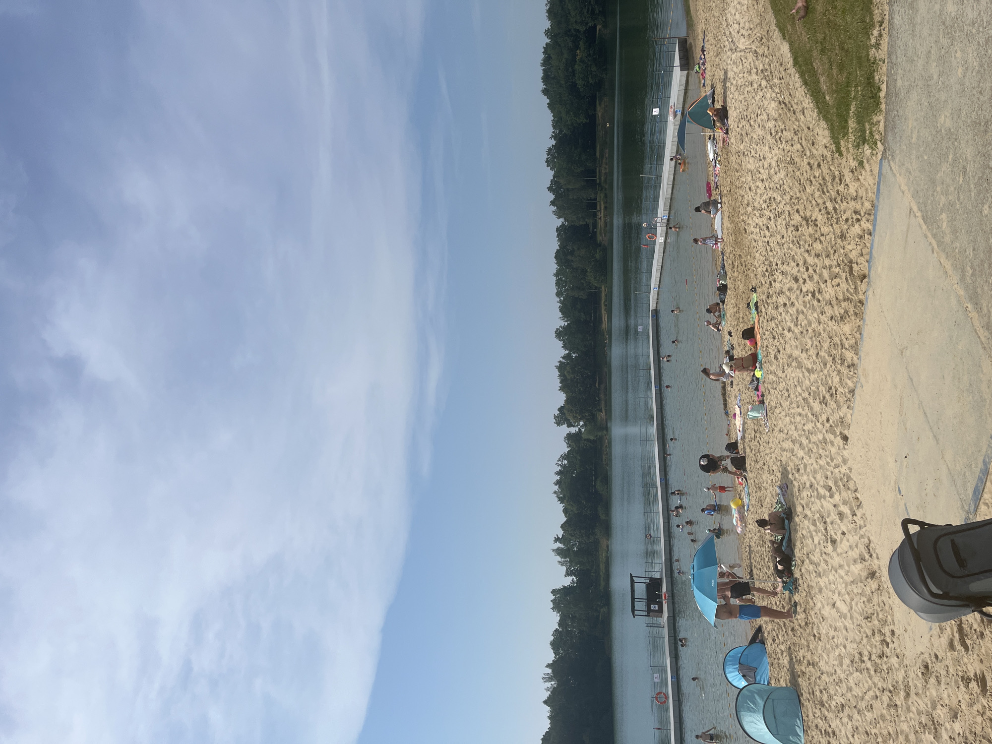 The image captures a sunny day at a crowded beach and swimming area. People are engaged in various leisure activities, including swimming, sunbathing, and relaxing under umbrellas. The scene is divided between a sandy beach with beachgoers and a large body of water, likely a lake or pool, where many are swimming and enjoying the water. Several blue umbrellas provide shade on the sand, and some people are sitting or lying on towels. In the water, there are numerous individuals, some swimming and others wading near the edge. A lifeguard stand is visible near the water's edge, suggesting a supervised recreational area. The background features a dense line of trees, indicating a natural or park-like setting. The sky is bright blue with some wispy white clouds, implying good weather. The overall atmosphere appears relaxed and joyful, typical of a summer day at a popular recreational spot. There is a dark gray stroller or cart in the foreground on the right side of the image. Some signs with white symbols on a red background are visible along the edge of the water.