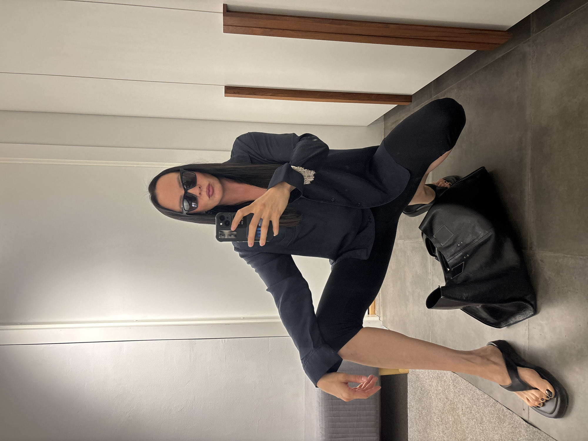 A person is posing in a mirror, possibly in a clothing store or dressing room. The person is wearing sunglasses, a dark-colored jacket or shirt, and dark leggings. They are holding a smartphone, presumably taking a selfie. A black handbag is on the floor next to them. They are wearing dark-colored flip-flops. The background appears to be a simple room with neutral-colored walls, accented by horizontal wooden trim. It is not possible to identify the location as Chaikovsky, Russia, without additional information or landmarks within the image.