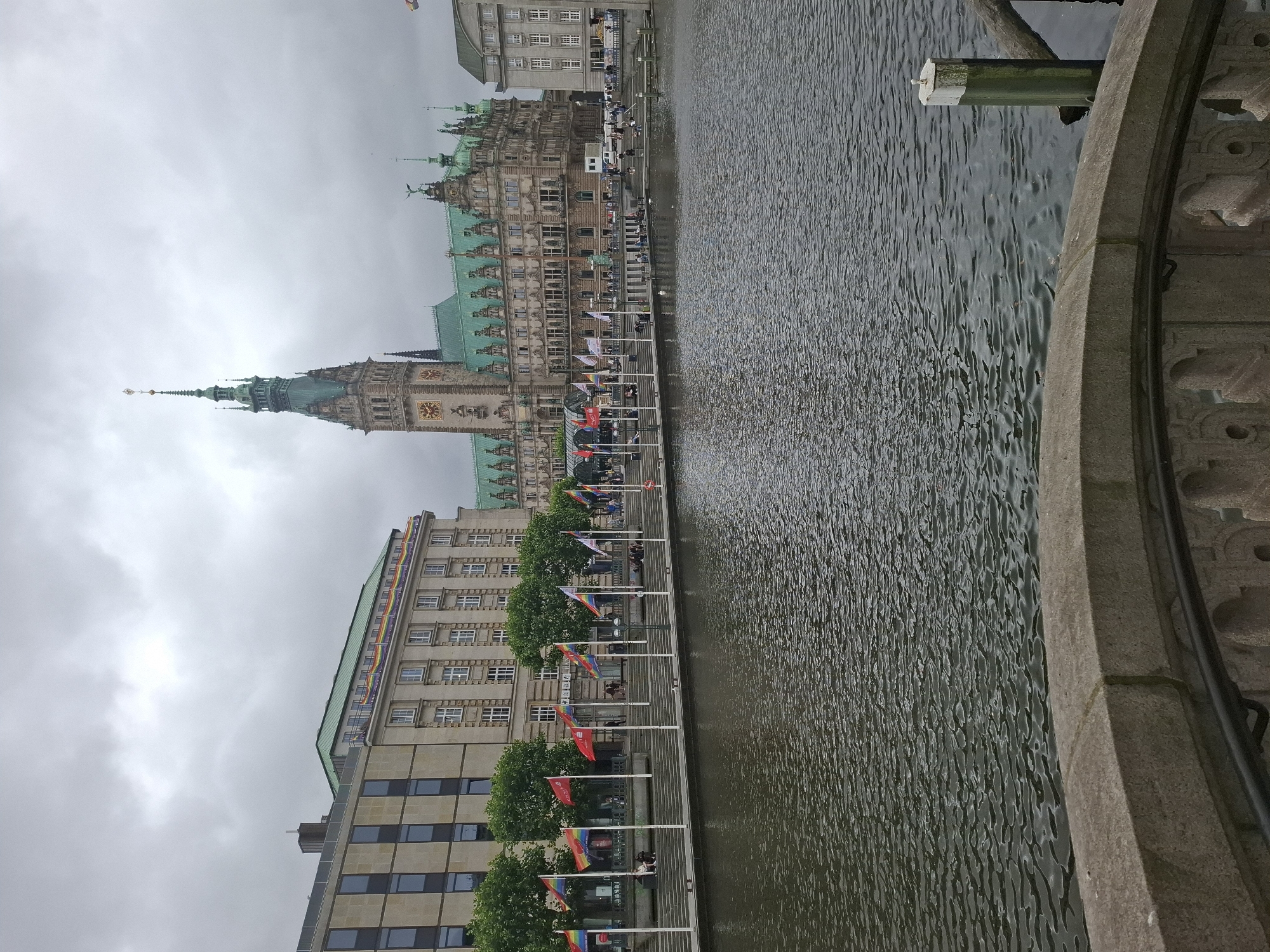The image shows a canal in Hamburg, Germany, with the Hamburg Rathaus (City Hall) in the background. The Rathaus is a large, ornate building with a tall spire. In front of the Rathaus, a canal reflects the cloudy sky. Along the canal, there are flags flying, including several rainbow flags, suggesting a Pride event. Several people are visible sitting on the steps leading down to the canal and walking along the promenade. The overall atmosphere is calm and overcast.
