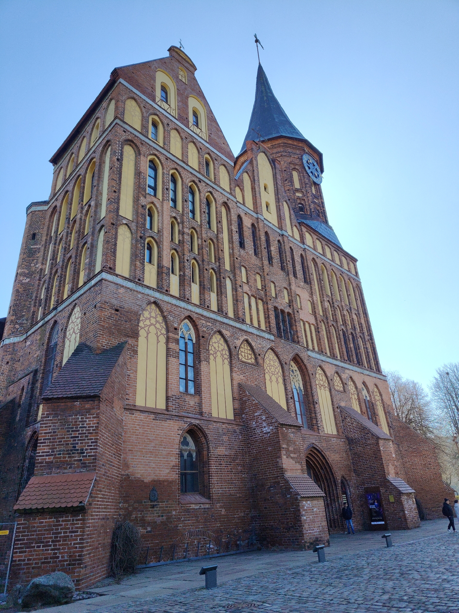The image shows a large brick building in the city of Unknown, Unknown. The building has a tall tower with a clock on it and a pointed spire. The architecture style has arched windows and yellow accents. There are people standing near the entrance of the building. The ground is paved with cobblestones. There are trees without leaves in the background. The sky is light blue.