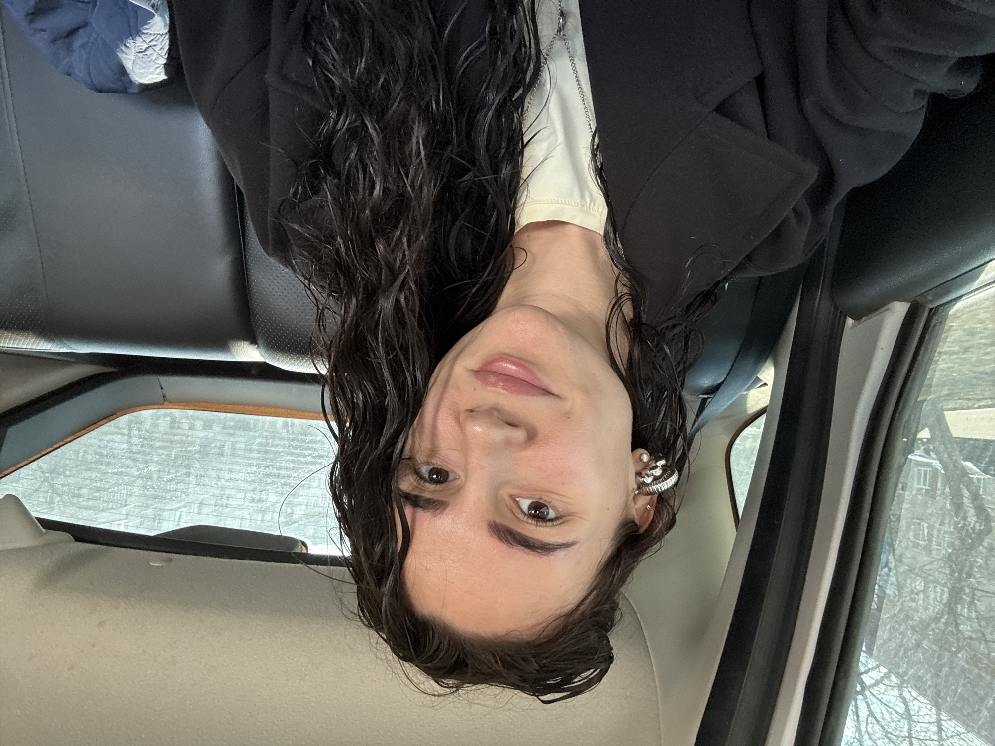 The image is a close-up selfie of a young woman, taken inside what appears to be a car. She is seen from the chest up, with long, dark, wavy hair that looks wet or styled with product. She has dark eyebrows and brown eyes, and her lips are naturally pink and slightly parted. She wears a light-colored top under a dark, possibly black, jacket or coat. Her left ear (viewer's right) is adorned with multiple silver-toned earrings, including a distinctive ear cuff. Her expression is neutral and calm.
The setting is the interior of a vehicle, with dark, possibly leather, seats visible behind and beside her. A blue, padded item of clothing is visible on the seat to her right. Through the car windows, a winter landscape is evident. The rear window shows a frosted or snow-covered view, and the side window reveals bare tree branches and a distant building, suggesting an urban or suburban environment during cold weather. The lighting is soft and diffused, indicating daytime, possibly overcast. There is no visible text. The overall scene depicts a casual moment in a car, consistent with winter conditions in Chaikovsky, Russia.