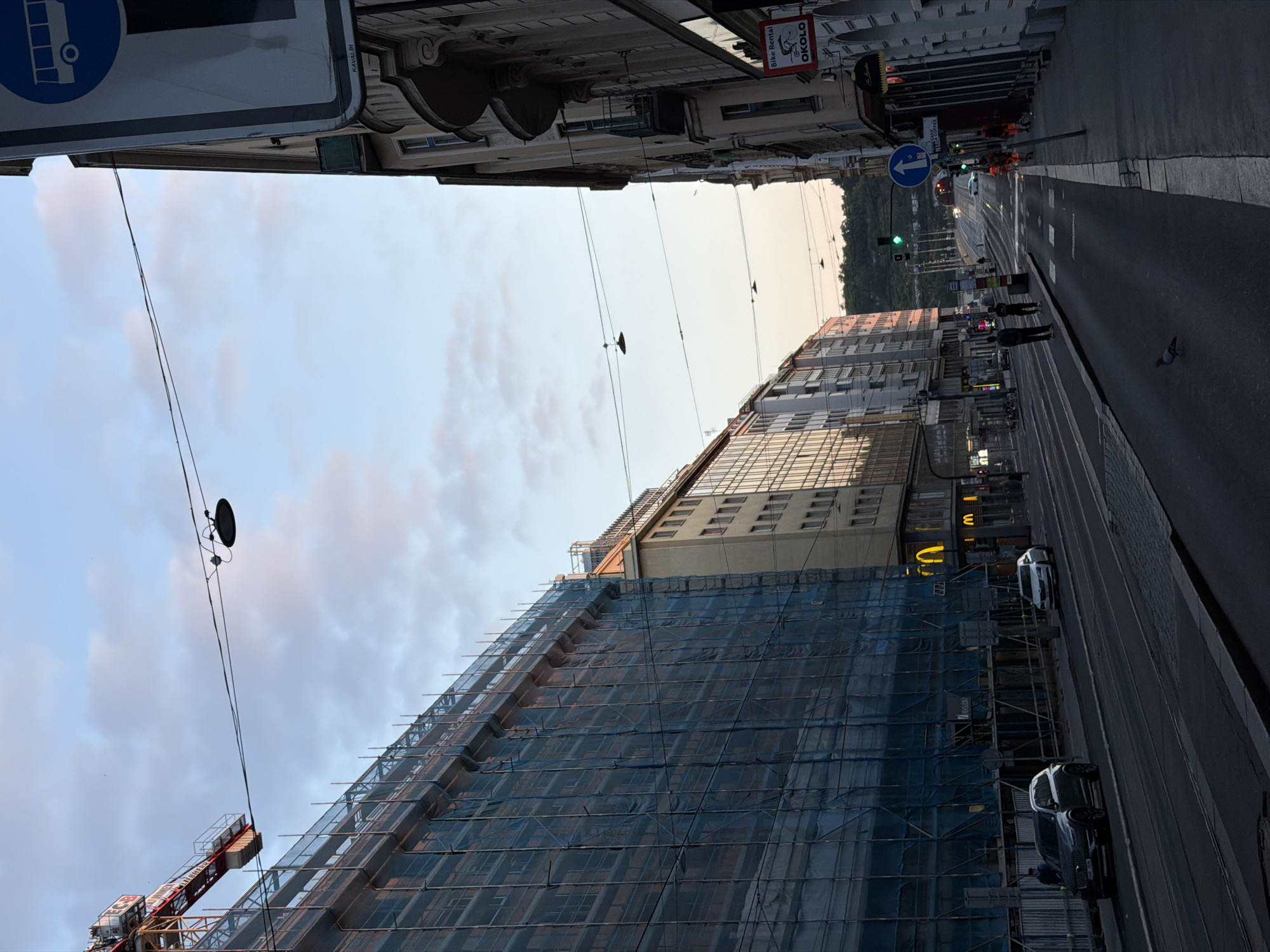 The image depicts a street scene in Prague, Czechia, likely in the late afternoon or early evening, as indicated by the warm lighting and the sky. On the left side of the street, a large building is under extensive scaffolding, covered with blue netting, suggesting ongoing construction or renovation. A red construction crane is visible above the scaffolding. Further down this side of the street, a McDonald's restaurant with illuminated golden arches is situated on the ground floor of a building.
The street itself is paved and appears to be a tram line, with visible tracks. Several cars are present, including a dark SUV parked near the construction site and a white car further down the street. Pedestrians are visible on the sidewalks and crossing the street. One person in an orange high-visibility vest is walking along the sidewalk on the right.
The right side of the street is lined with buildings featuring traditional architecture, with balconies and ornate details. A sign for "Bike Rental OKOLO" is affixed to one of these buildings. Traffic lights are visible, with one showing green. Overhead wires for streetlights or utilities are strung across the street. The background shows a distant hill with greenery, and a tunnel entrance is visible in the distance. The sky is a mix of pale blue and soft pinkish-white clouds. A pigeon is walking on the street in the foreground.