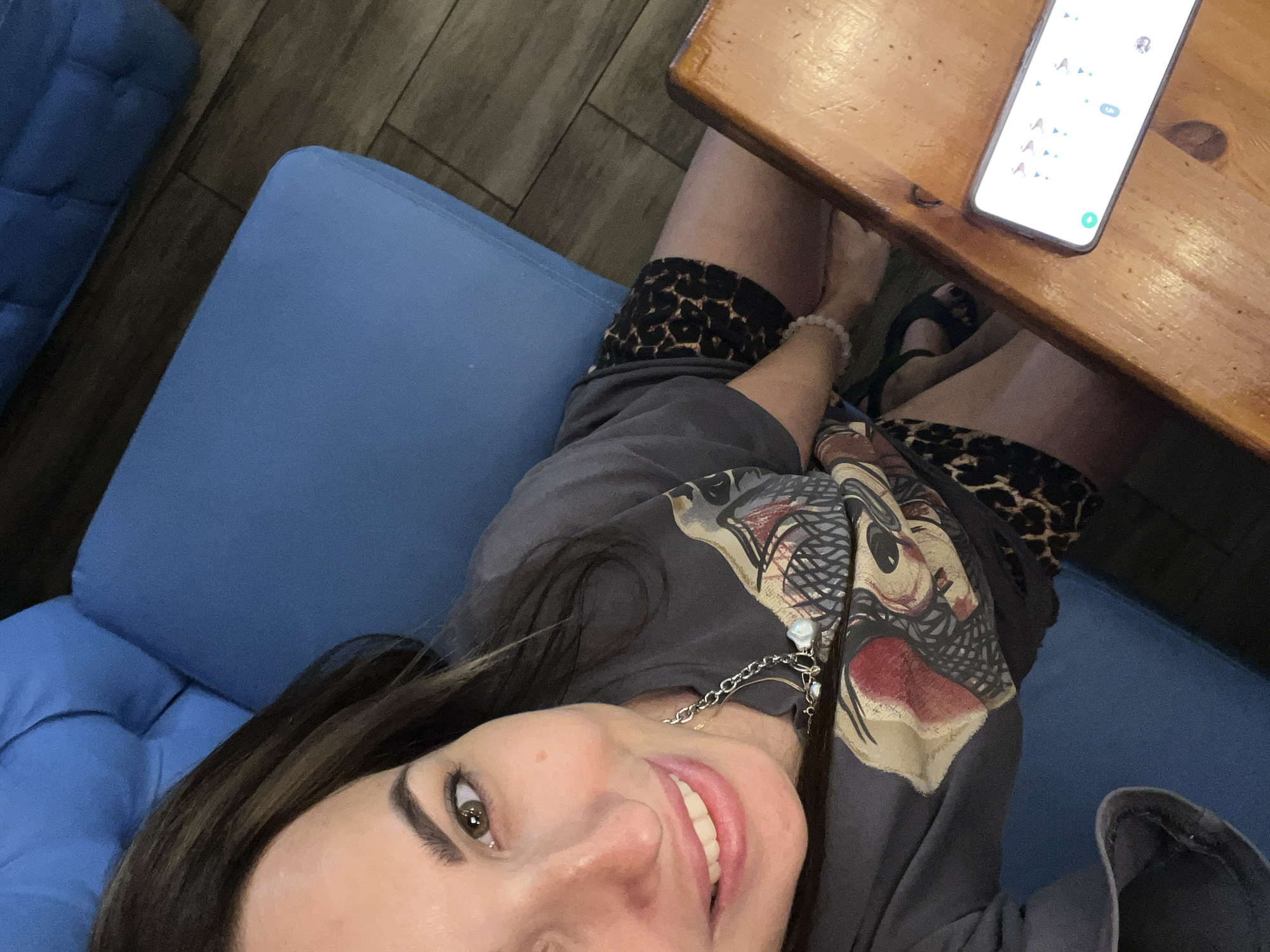 A woman with dark brown hair, wearing a grey t-shirt with a graphic print and leopard-print shorts, sits on a blue cushioned bench in an indoor setting. She is leaning forward, smiling at the camera, with a silver chain necklace around her neck. Her right hand rests on a wooden table where a smartphone is placed. The background consists of more blue cushioned furniture and a wooden floor. The scene is lit by indoor lighting, casting soft shadows.