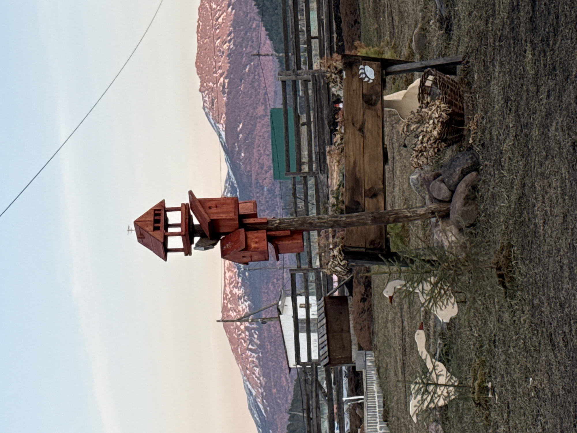 This outdoor shot depicts a birdhouse resembling a multi-tiered structure, atop a wooden post. Adjacent to it, wooden geese stand amidst dense greenery, adding a decorative touch. In the background, a wooden fence separates the foreground from a landscape featuring mountains. The image was captured in Tyulyuk, Russia.