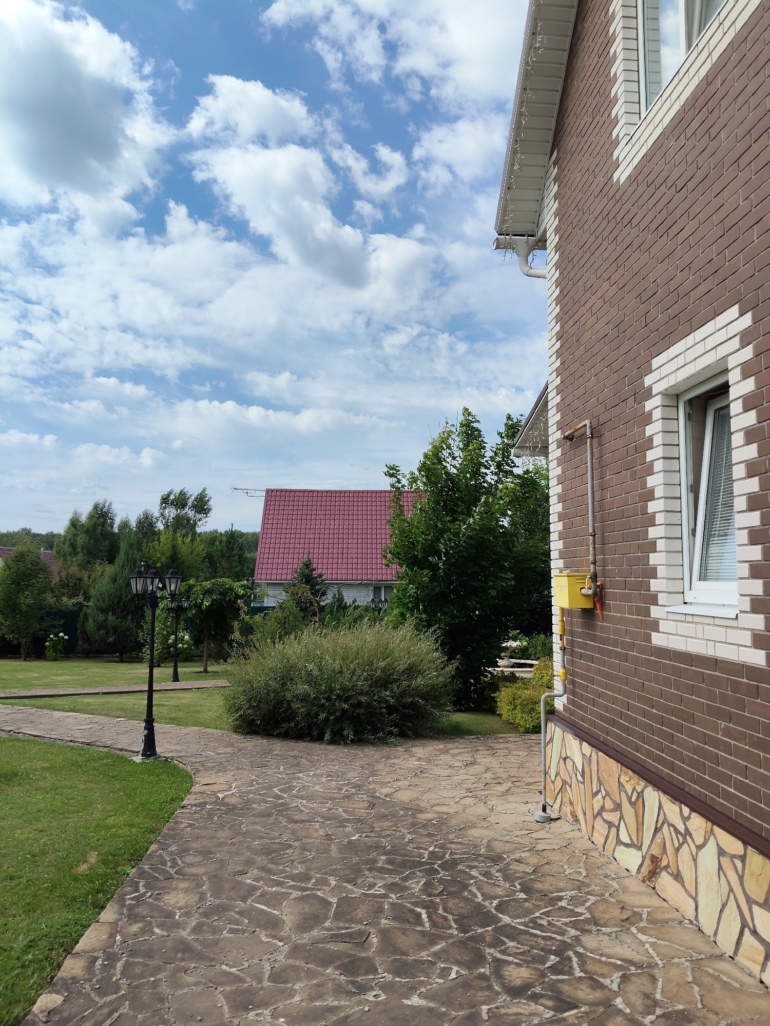 The image depicts a side view of a two-story house made of brown and white brick, with a prominent white-framed window on the right side. A yellow gas meter box is mounted on the brick wall. The house has a white gutter system and decorative lights strung along the eaves. A paved pathway made of irregular stone slabs leads from the foreground towards the house and curves to the left. To the left of the pathway, a lush green lawn is visible, with a black lamppost standing on the grass. In the background, across the lawn and a dense bush, a portion of another house with a red roof is visible, surrounded by numerous trees. The sky above is a bright blue with scattered white clouds. The overall scene appears to be a residential area, possibly in Bogdanovka, Russia, given the architectural style and vegetation. There are no people or animals present in the image.