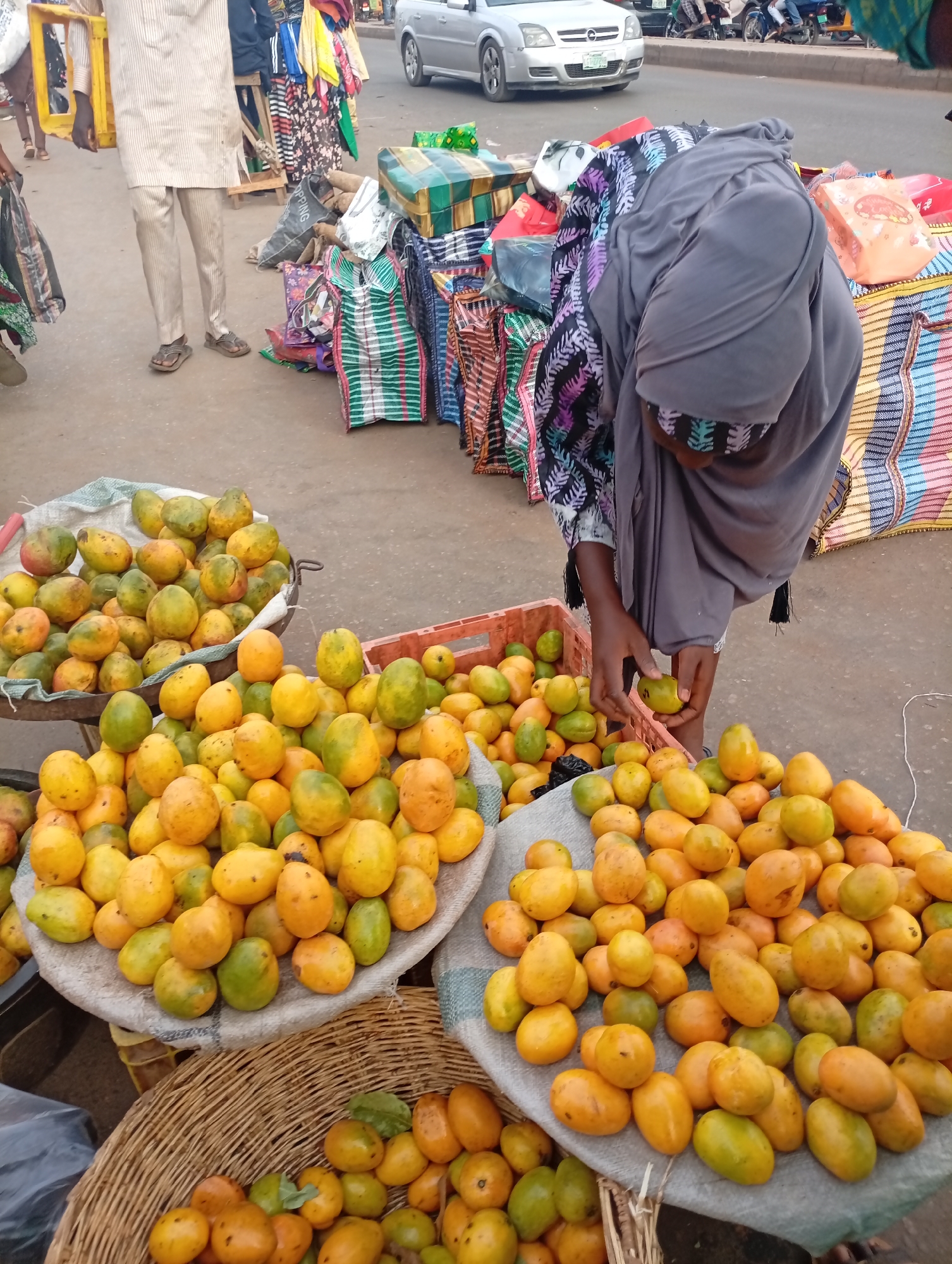 The image shows an outdoor market scene in Sabon Tasha, Nigeria. A woman wearing a grey hijab is sorting mangoes, organizing them between a red plastic crate and a pile resting on a grey cloth. Several baskets and metal trays containing more mangoes are visible. In the background, a man is standing in a striped shirt, and colorful bags and goods are piled up. A silver car drives along the street on the right side of the image.