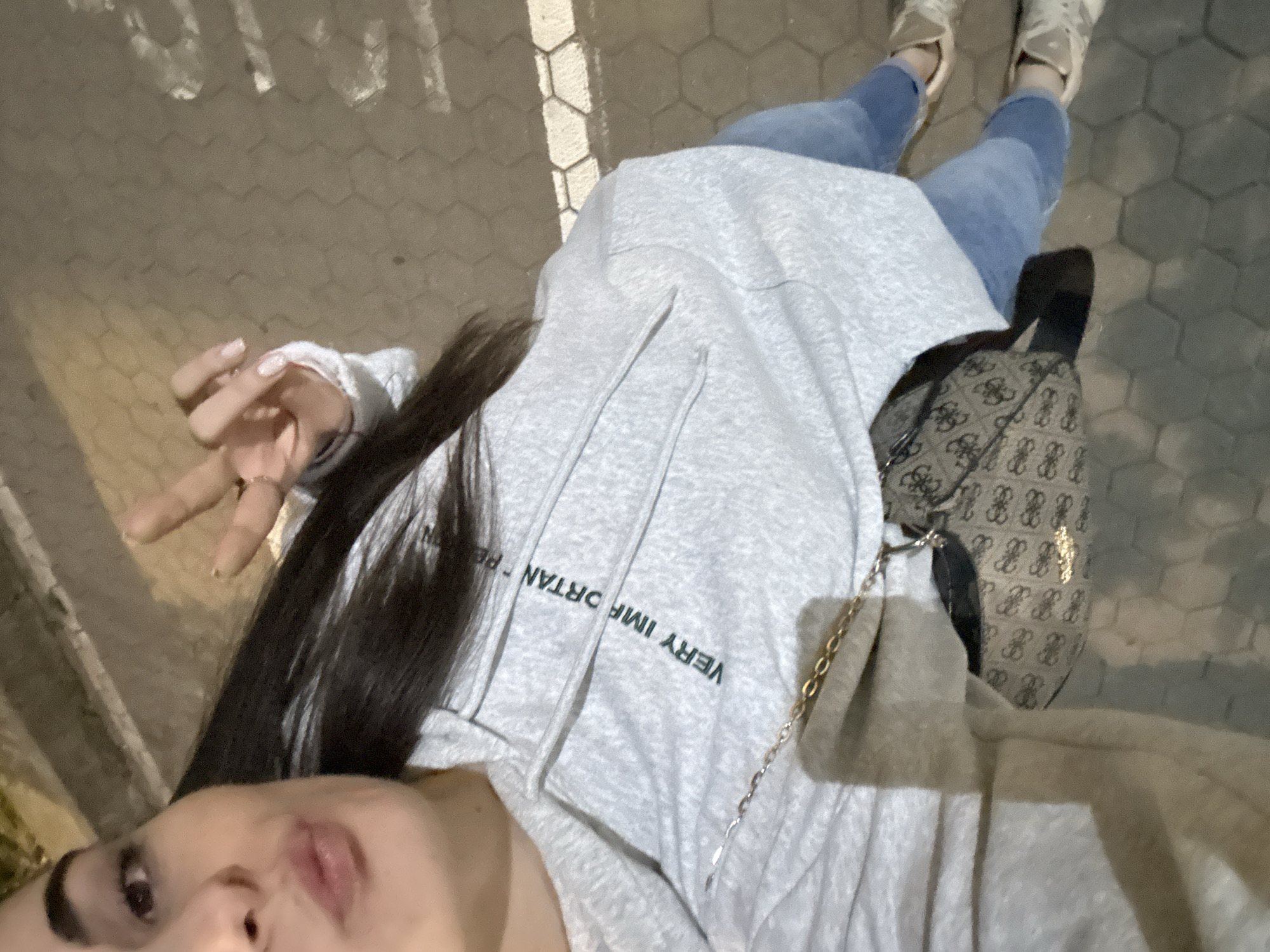 An overhead shot shows a person from the chest down in a city environment. The person is wearing a light grey hooded sweatshirt with the text "VERY IMPORTANT PERSON" printed in dark green letters. A small, patterned handbag with a gold chain strap is visible, resting on the person's lap. The person is also wearing light blue jeans and white sneakers. They are standing on a hexagonal-patterned pavement. The background appears to be a walkway with some lighting. The person's left hand is raised, with two fingers extended in a peace sign.