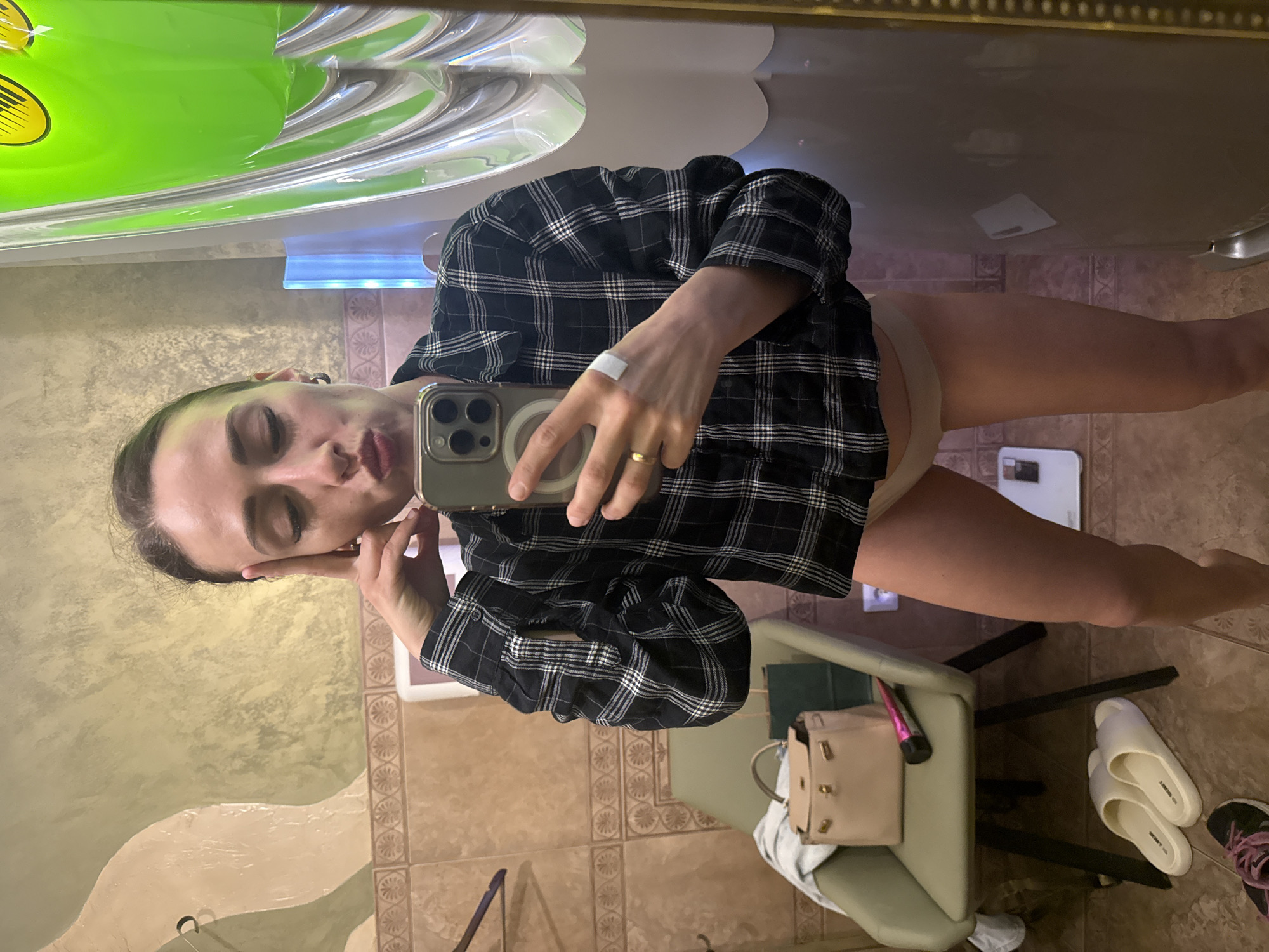 The image is a mirror selfie taken by a woman in an indoor setting, likely a changing room or bathroom, possibly within a solarium or spa in Saint Petersburg, Russia.
The central subject is a woman, holding a smartphone (resembling an iPhone with a MagSafe ring on its case) with her right hand to capture her reflection. She is wearing a black and white plaid shirt, unbuttoned, over light beige briefs or underwear. A gold-colored ring is on her right ring finger, and a white bandage or plaster is visible on her right wrist. Her left hand touches her face, which has a relaxed expression with eyes partially closed and lips slightly pursed. Her dark hair is pulled back.
The room features mottled yellow and beige textured walls and tiled flooring with decorative brown and beige patterns. In the mirror's reflection, behind the woman, a prominent bright green object with yellow accents is visible, resembling part of a tanning bed or similar equipment, along with white, curved, reflective panels and a blue light source.
On the floor, near a light-colored chair with a green/beige seat and dark frame, a beige/pink handbag with gold hardware is placed. A slender pink object, possibly a makeup brush, rests next to the bag. A pair of white slip-on sandals, with the partial text "HO BOGOT" or "HO SOGOT" visible on one, and a black and pink athletic shoe are also on the floor. Two small white rectangular objects are seen in the background, one on the floor and another on a shelf or surface. The overall scene suggests a private and casual moment.