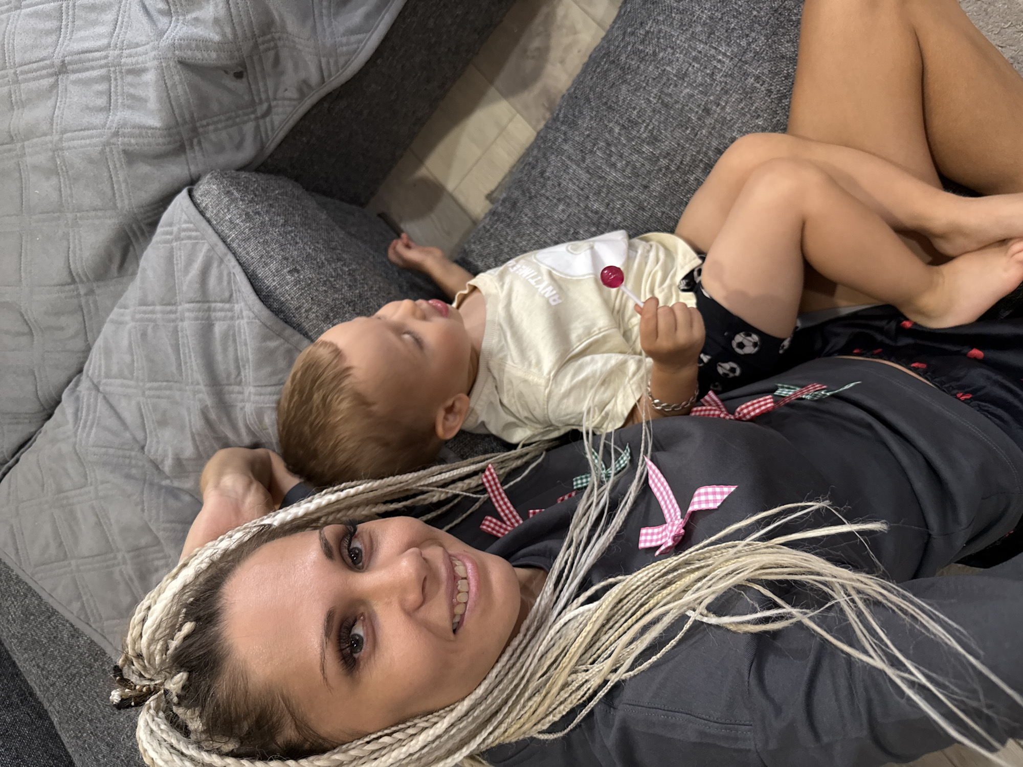 A woman with blonde braided hair and a grey t-shirt sits on a grey couch, holding a young child on her lap. The child wears a cream-colored t-shirt and black shorts with soccer ball patterns. The child is holding a red lollipop. The woman is smiling and looking at the camera. The scene is likely indoors, possibly in a living room. The couch appears to be made of fabric.