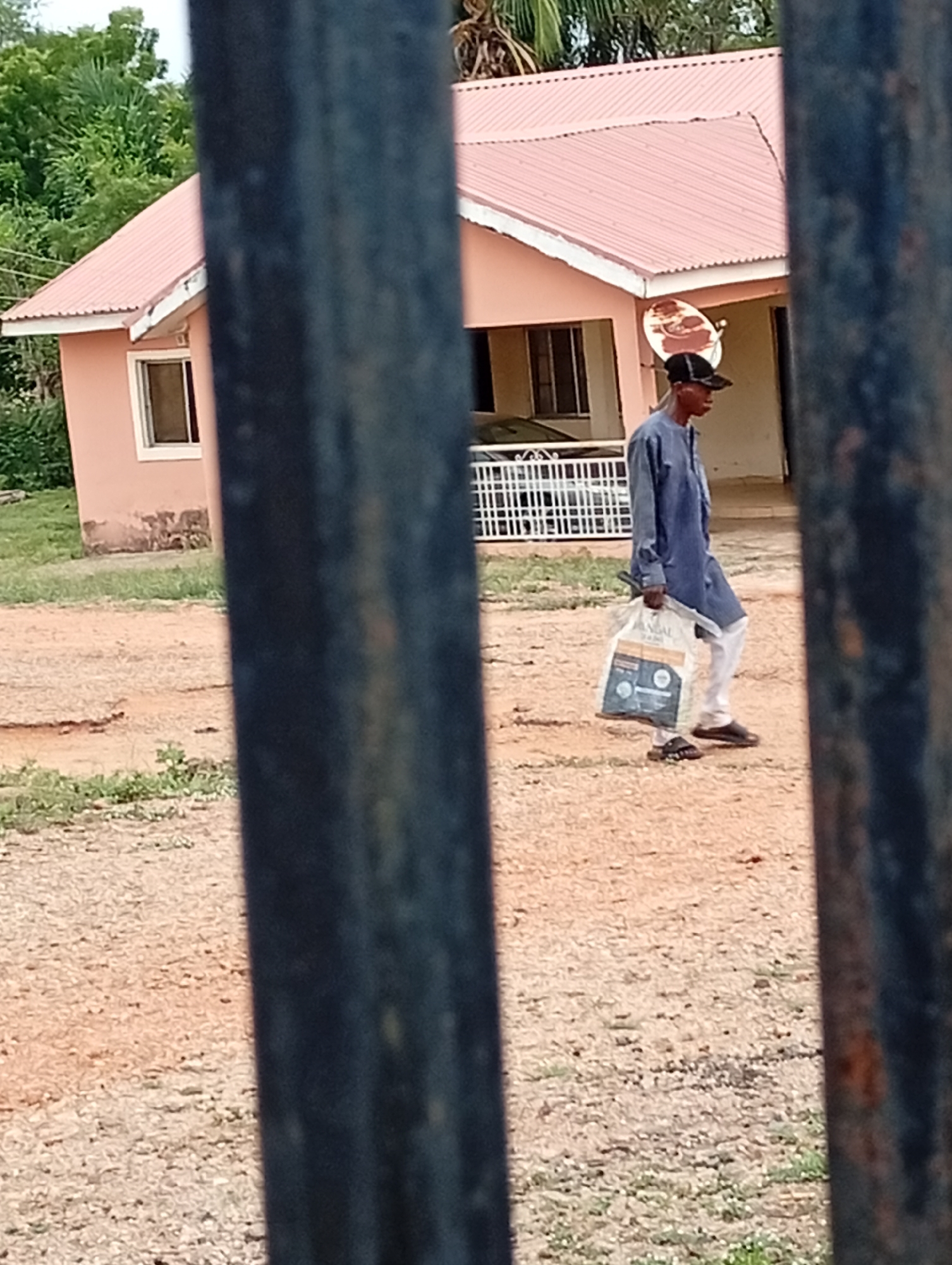 The scene is in Adankolo, Nigeria. Through a metal fence, a single-story pink building with a reddish roof is visible. A satellite dish is mounted on the building's exterior. A man wearing a dark cap, blue top, light trousers, and sandals walks across a dirt-covered yard, carrying a white shopping bag. A vehicle is behind a short white picket fence in front of the building. Grass and trees are sparsely present in the background.