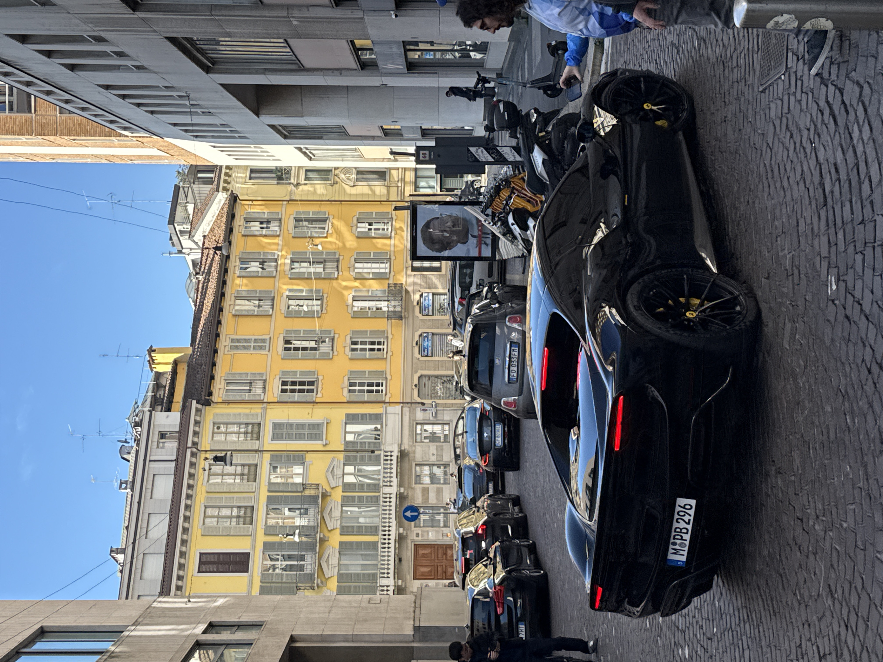 The image depicts an urban street scene in Milan, Italy, under a clear blue sky. In the foreground, a black Ferrari sports car, bearing German license plate M•PB 296, is positioned on a cobblestone street, its rear facing the viewer with red taillights illuminated. Yellow brake calipers are visible through its black multi-spoke wheels.
To the right of the Ferrari, a man with dark, curly hair and glasses, wearing a blue and white patterned jacket over a dark shirt and grey pants, is partially visible, holding a black smartphone to photograph or video the car. A cylindrical grey bollard is present at the extreme right.
Further down the cobblestone street, a line of vehicles is stopped or parked. These include a black BMW (partially visible license plate ending in 7RT), a dark SUV, and a grey Fiat 500 with Italian license plate FD 033FM. On the left side of the street, a man dressed in dark attire walks away from the camera, focused on his mobile phone.
The architectural environment consists of a mix of historic and modern structures. On the left and directly behind the parked cars are multi-story buildings with bright yellow facades, featuring numerous windows with light grey shutters, some open, some closed. Balconies with ornate railings and decorative elements are present. Ground-floor establishments in these yellow buildings display "Borsa Affari" signage. A blue circular street sign with a white upward arrow indicating a one-way street is visible near a large wooden door. On the right, contemporary, grey concrete or stone buildings with prominent vertical window designs and large ground-level windows frame the street. Rooftops are visible in the background, adorned with various antennas and chimneys.
Motorcycles and an electric scooter are parked along the right-side sidewalk. A large outdoor digital display panel, featuring a dark-toned image of a person's profile, stands further down the street on the right.