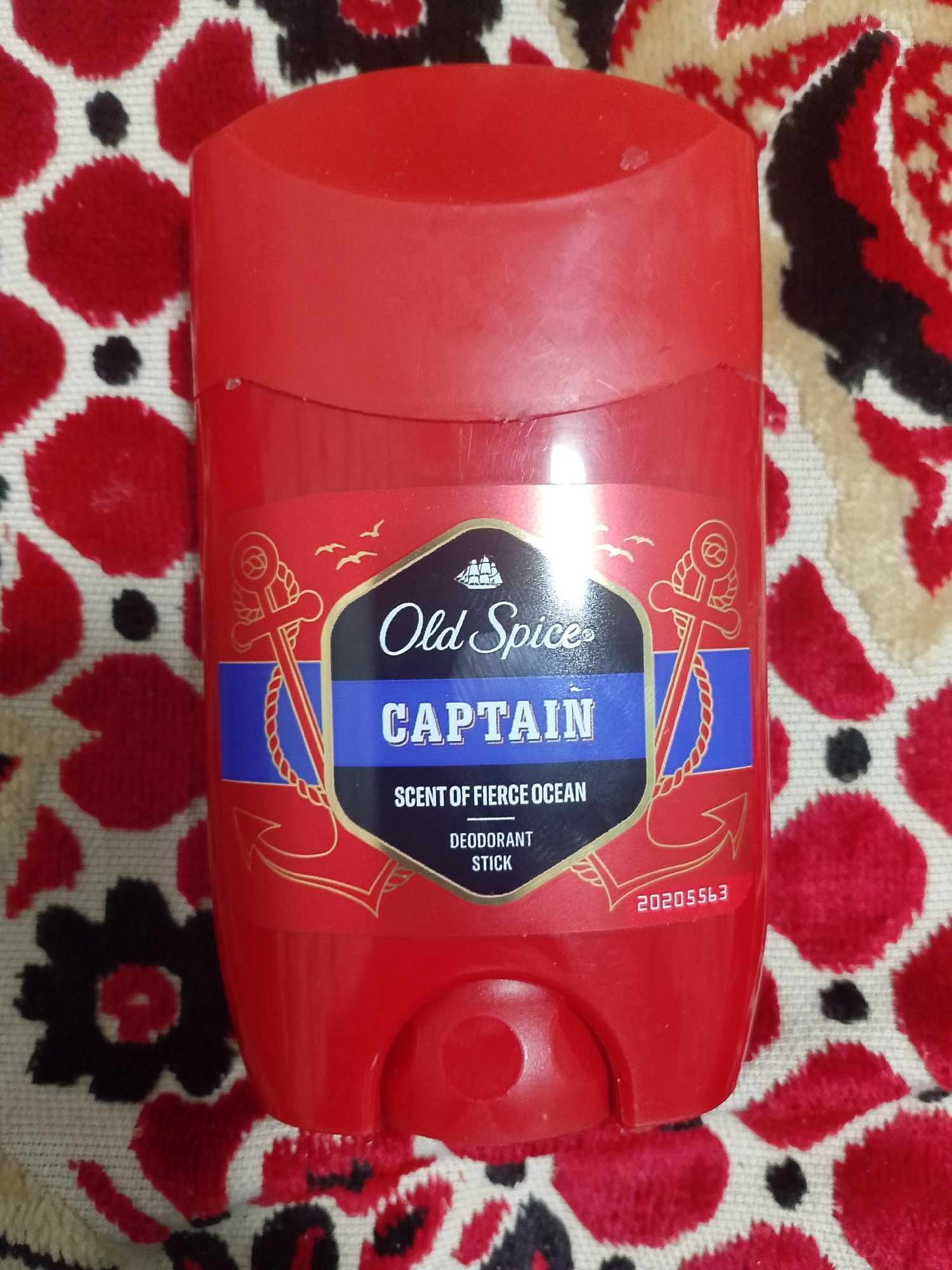 A red Old Spice Captain deodorant stick is shown lying on a red and black floral patterned fabric. The deodorant has a cap and a label with "Old Spice CAPTAIN SCENT OF FIERCE OCEAN DEODORANT STICK" printed on it. A number, "20205563", is visible on the bottom of the deodorant. The image does not contain any information about the city of Asino, Russia.