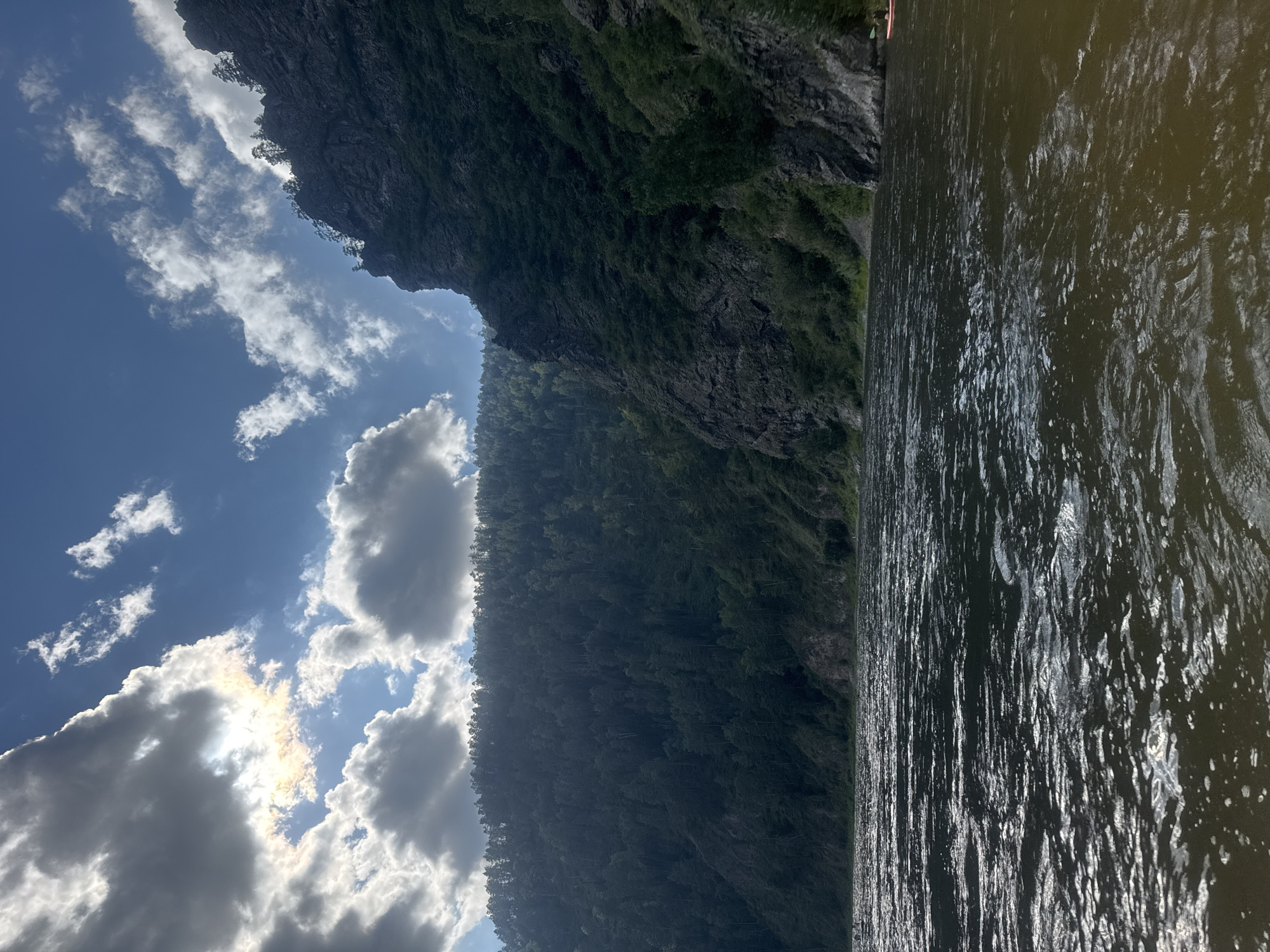 The image depicts a wide, choppy river flowing past steep, tree-covered mountainsides under a dramatic sky filled with sunlit clouds. The water in the foreground is dark and reflective, with whitecaps indicating movement. On the left, a dense forest covers the slopes of a mountain that recedes into the distance. On the right, a sheer rock face rises abruptly from the riverbank, also covered in patches of vegetation and trees. The sky is a vibrant blue, with large, white cumulus clouds partially obscuring the sun, creating rays of light that pierce through. The overall scene suggests a wild, natural landscape. The location is identified as Ust-Mana, Russia, a settlement in the Krasnoyarsk Krai region, known for its scenic natural beauty and river activities. There is a small, distant red boat with a paddle visible on the right side of the river.