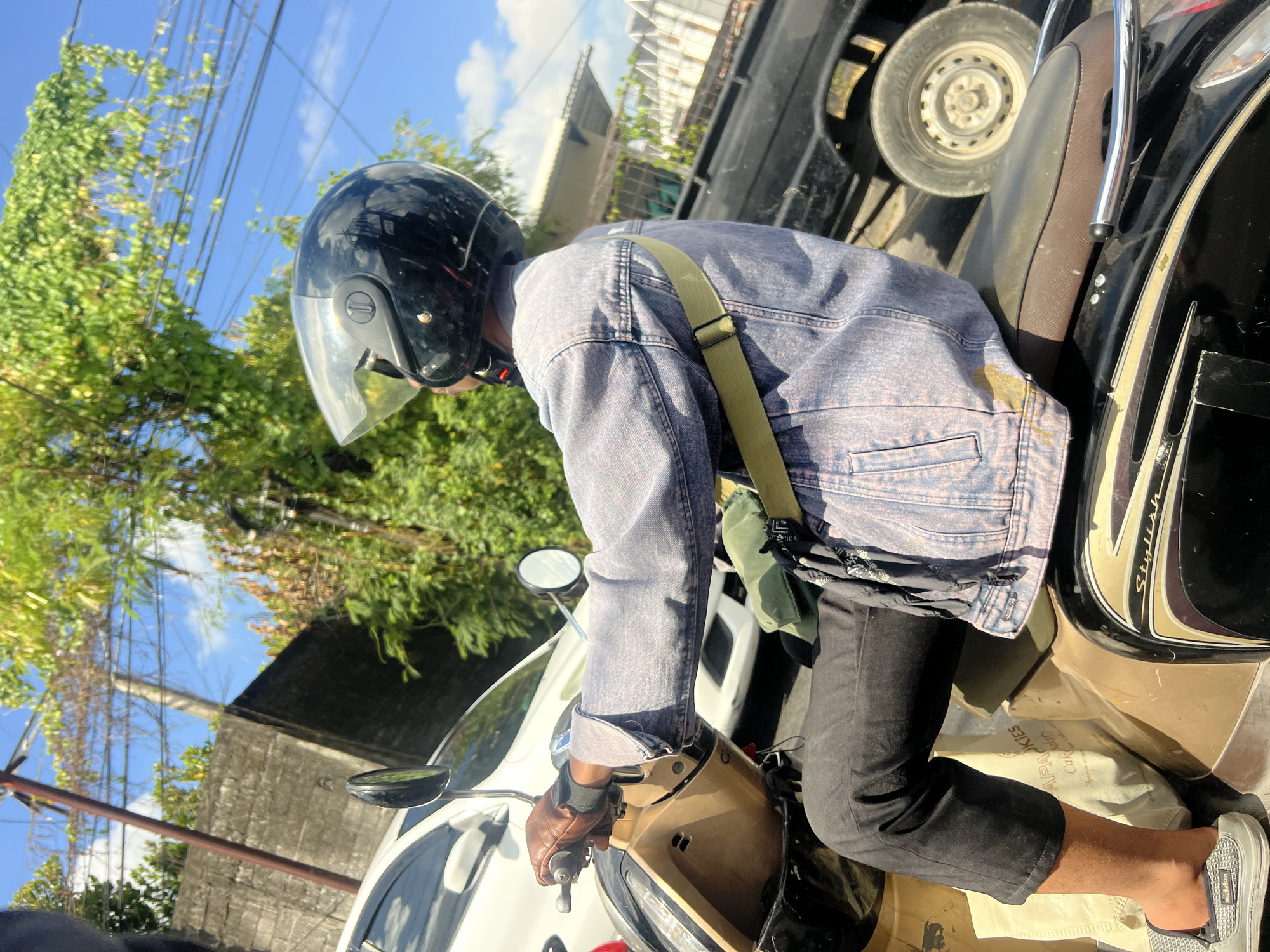 A medium-angle shot captures a person riding a scooter on a street, with a white car to their left and a dark truck to their right. The rider, wearing a blue denim jacket, black pants, a black helmet with a clear visor, and brown gloves, is holding onto the scooter's handlebars. A green bag is slung across their shoulder, and a beige paper bag is visible on the floorboard. The background features a concrete wall, lush green trees, power lines, and a clear blue sky with scattered clouds, indicating a sunny day. The scene appears to be in Denpasar, Indonesia, likely a residential or commercial area given the mix of vehicles, buildings, and vegetation.