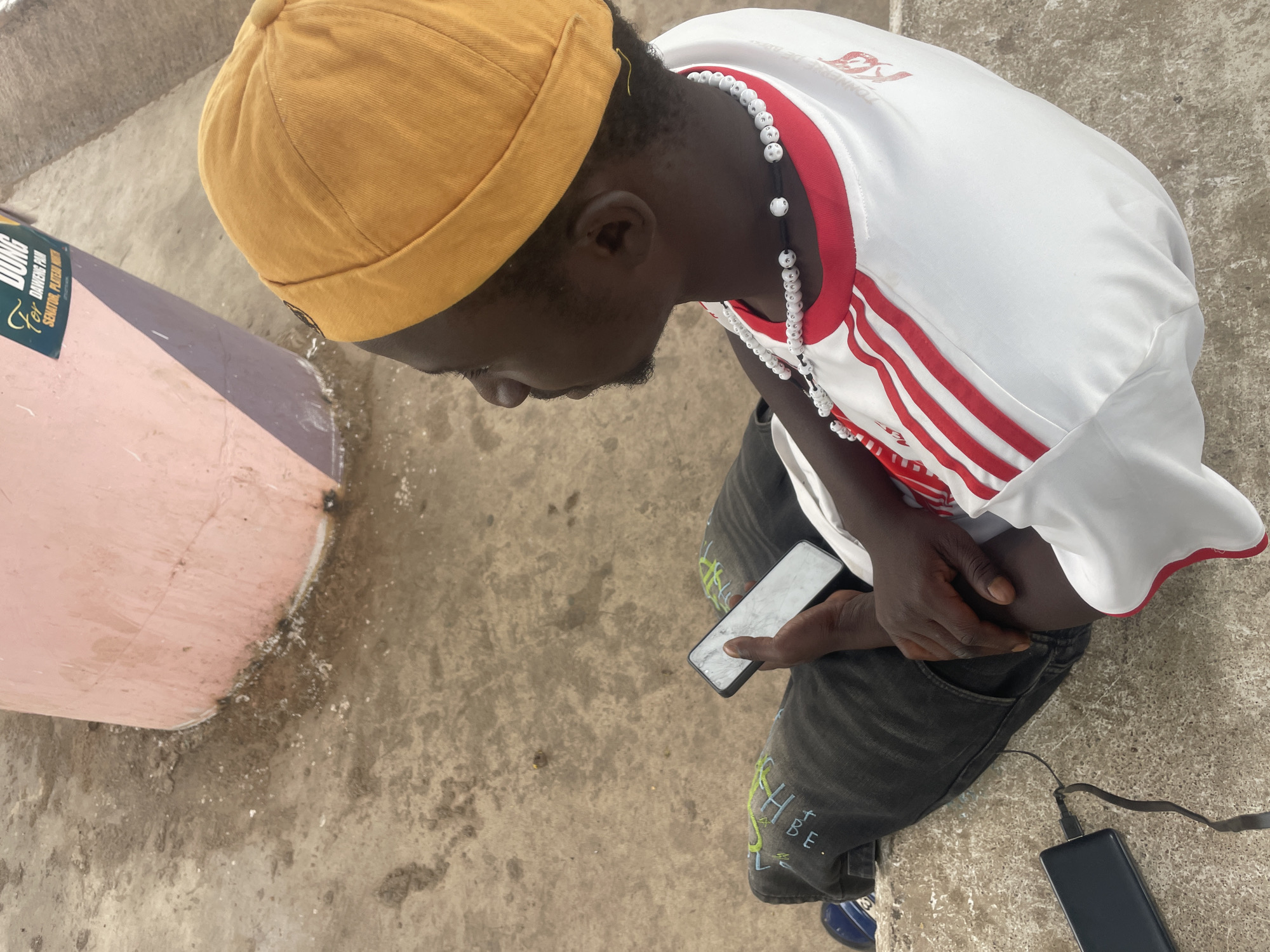 The image shows a young Black man from an overhead perspective, using a smartphone. He is wearing a mustard-colored hat, a white jersey with red stripes on the shoulders, and a beaded necklace. His black jeans have light-colored graffiti designs. He is sitting on a concrete ledge, with a portable power bank connected to his phone via a black cable. A cylindrical object painted in shades of pink and lavender is visible to his left. The location is inferred to be Jos, Nigeria, based on signage on the cylindrical object. The scene appears to be outdoors, with a concrete surface serving as the ground.