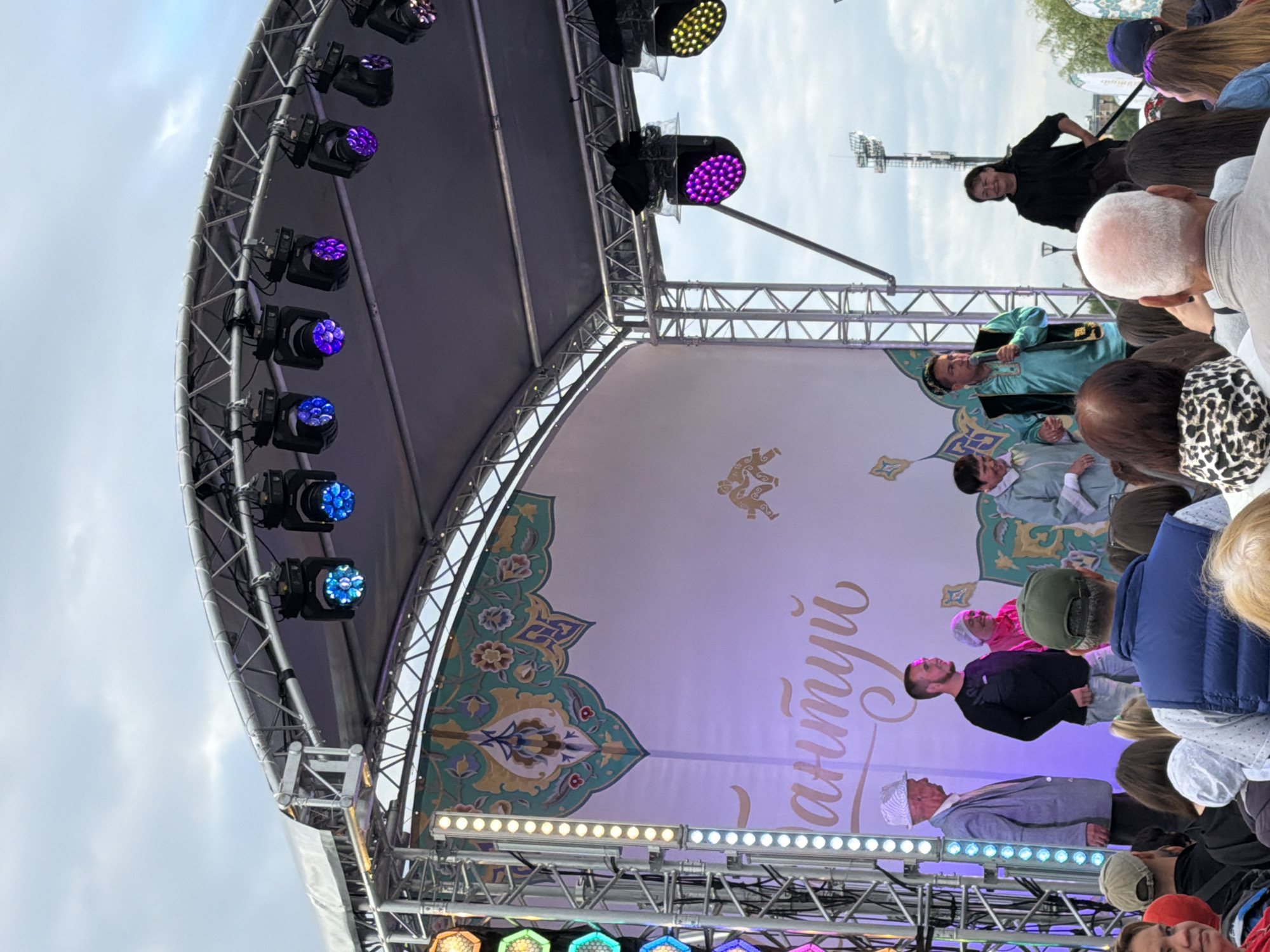 The image shows an outdoor stage event with a crowd of people in the foreground watching performers on stage. The stage is set up with a large banner in the background featuring ornate, colorful designs and what appears to be text in a script. There are several stage lights suspended above the stage, some emitting blue and purple light. On stage, several individuals are visible. From left to right, an older man in a light-colored suit and hat stands with his back to the camera. Next to him, a man in a black long-sleeved shirt and jeans smiles. Behind them, a woman in a pink headscarf and jacket is visible. Further to the right, a man in a turquoise traditional garment with intricate gold embroidery is speaking into a microphone. To his right, a woman in a dark shirt and pants stands. The audience in the foreground consists of many individuals, some with their backs to the camera, suggesting they are facing the stage. The overall atmosphere appears to be festive, possibly a cultural or community event. The backdrop suggests an overcast or cloudy sky.