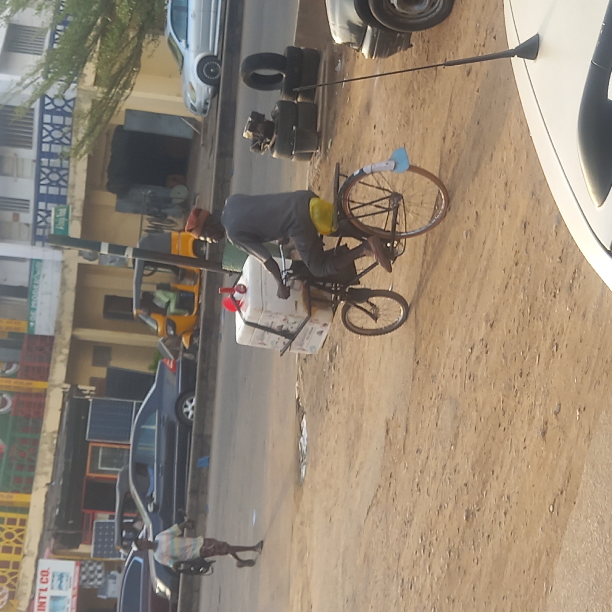 The image shows a street scene in what appears to be Dadi Riba, Nigeria. A man is riding a bicycle outfitted with a large white container, possibly for carrying water or some other commodity for sale. The bicycle has two wheels. He is wearing a gray shirt and an orange head covering. To the left, there is a yellow three-wheeled vehicle and several parked cars. A man is walking along the side of the road. In the background, there are buildings with shops and living spaces. There are stacks of tires beside a car, and another car parked next to it. The ground is a mix of dirt and paved surfaces.