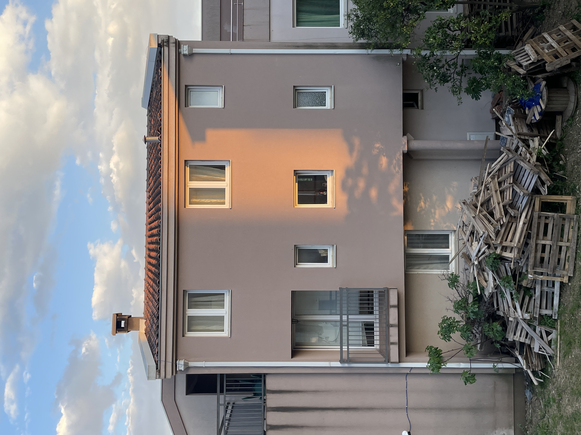 The image shows a multi-story building in Bar, Montenegro. The building is light brown with a red tile roof and several windows of varying sizes. A stack of wooden pallets is piled against the side of the building, partially obscuring a tree. The sky is visible with clouds. The building has a balcony with metal railings. The facade is illuminated by sunlight.
