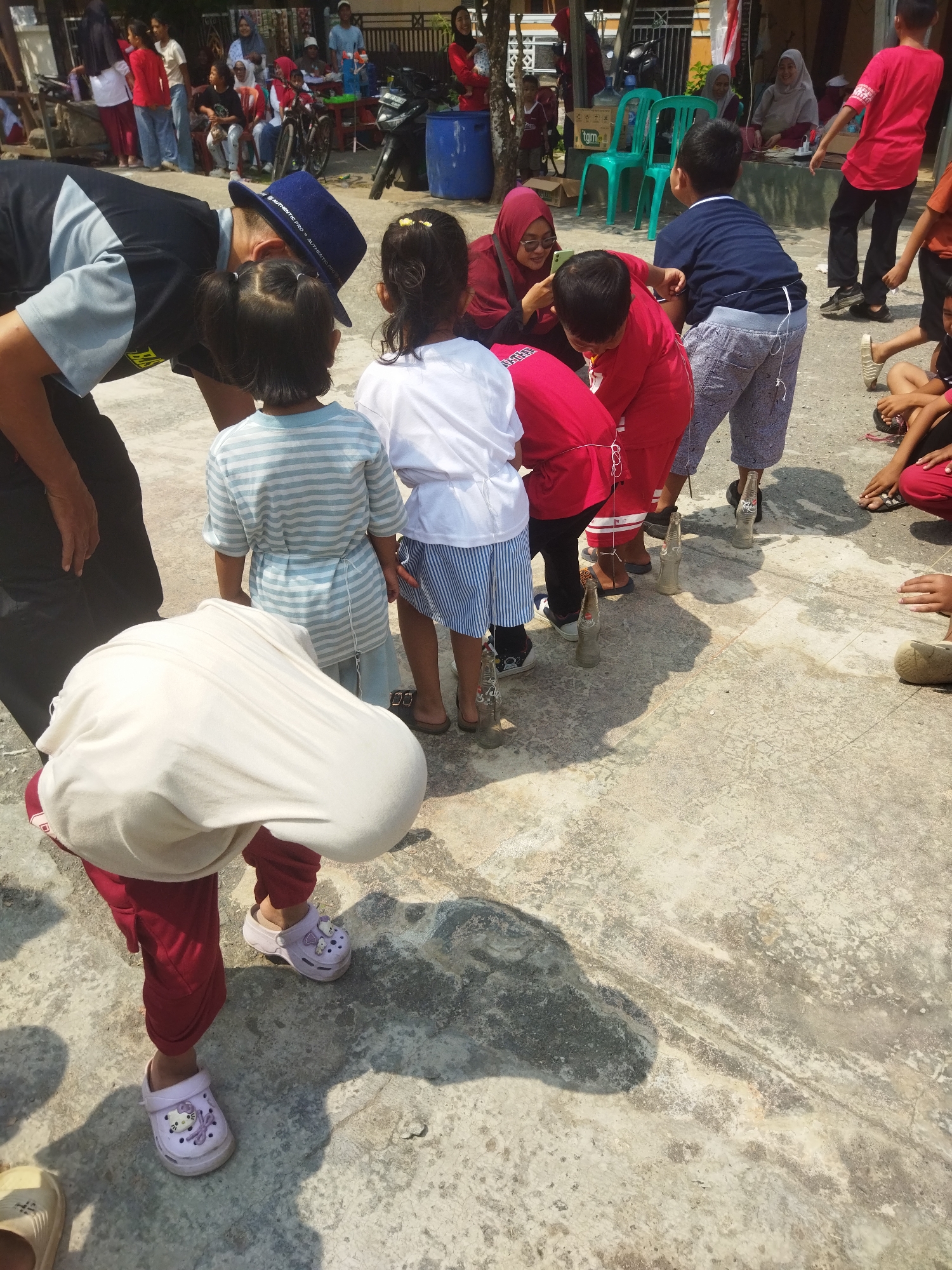 In Banjar, Indonesia, a group of children and adults are gathered on a concrete surface, possibly a yard or public space, under bright sunlight. The activity appears to be some form of game involving bottles and string. Several children, some wearing colorful clothing and others in more muted tones, are lined up. They are connected by strings that run from their waists to bottles placed on the ground in front of them, suggesting they are participating in a race or challenge where they must move the bottles using the strings.
One child in the foreground is bent over, wearing a white oversized garment and red pants, with purple sandals. Behind this child, another child in a blue and white striped shirt is also participating. In the background, an adult wearing a blue hat and black and gray shirt is observing the children, seemingly guiding or supervising. Other adults and children are present in the periphery, some watching the game and others engaged in their own activities. The environment includes a motorcycle, plastic chairs, and building structures in the background, indicating an outdoor setting in a populated area. The ground is textured concrete with visible cracks and shadows cast by the people and objects.