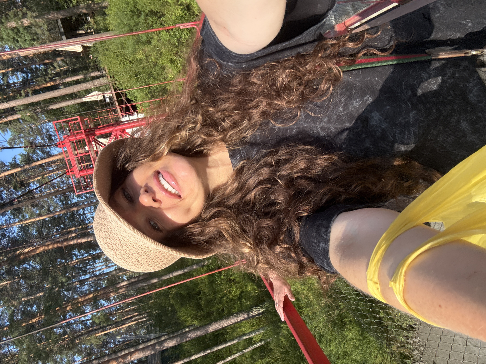 The image depicts a woman in a straw hat and a dark t-shirt, taking a selfie outdoors. She has long, curly brown hair and is smiling, sticking out her tongue slightly. She is holding a yellow plastic bag in her left arm, which is extended towards the camera. Her right hand is resting on a red railing. In the background, there are tall pine trees, green foliage, and a red ski lift structure. The scene appears to be a recreational area, possibly a park or resort, bathed in sunlight. The location is identified as Barangol, Russia.