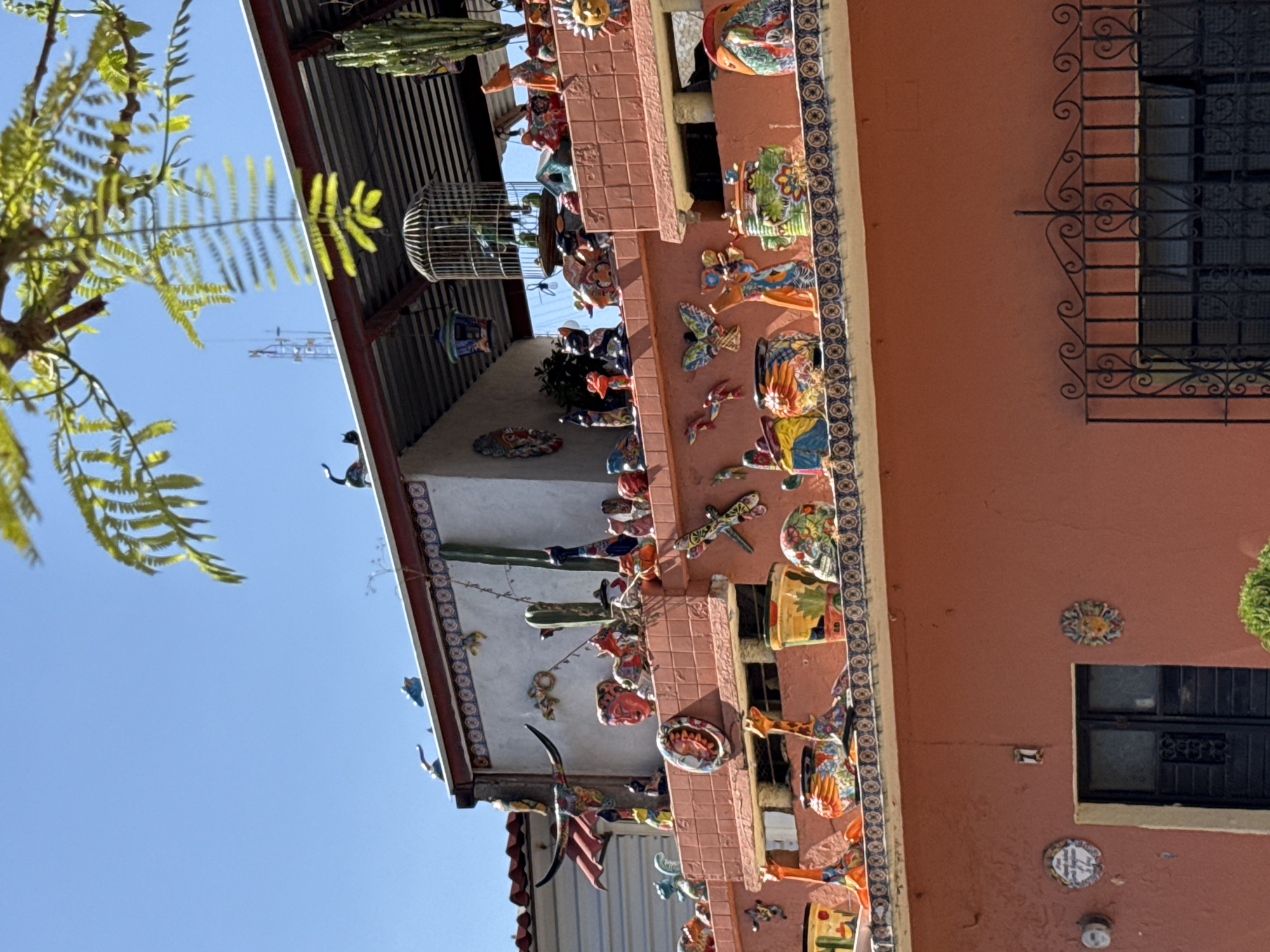 The image shows a section of a building exterior in Querétaro, Mexico. The building has a terracotta colored wall adorned with numerous, colorful ceramic decorations, including animals, suns, and geometric patterns. The building also features a decorative tiled trim along the edge. There are barred windows, one with a decorative wrought iron design. The roofline is visible, with a corrugated metal awning. A tree with green leaves is in the foreground. The sky is clear and blue.