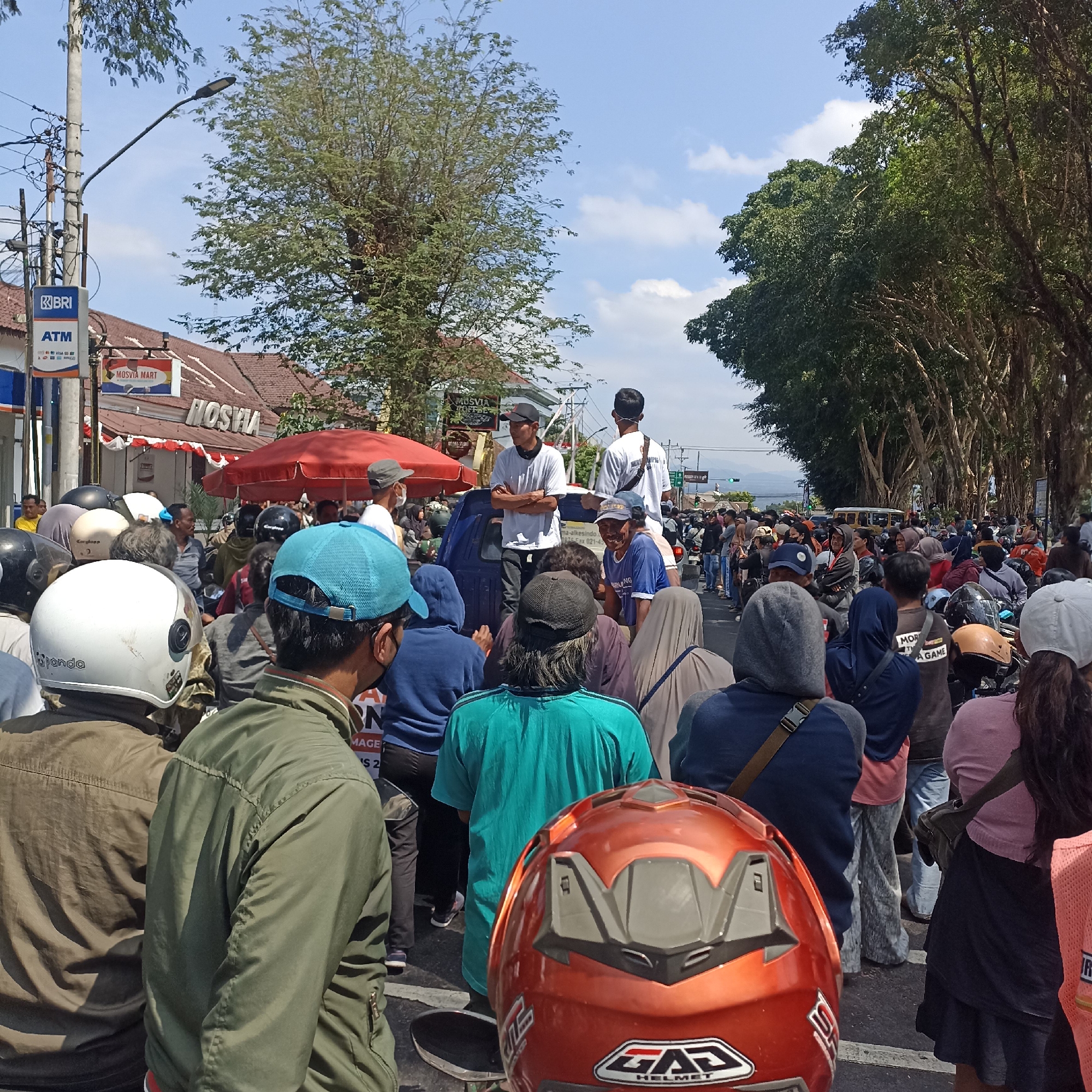 The image depicts a crowded street scene in Magelang, Indonesia, likely on a sunny day with scattered clouds. A dense group of people, many on motorcycles, are gathered. In the foreground, several individuals are visible from behind, wearing helmets and jackets. A prominent orange motorcycle helmet with "GAZ HELMET" branding is in the lower center.
Further back, a blue pick-up truck is positioned, with several people standing on its bed, including two men in white t-shirts. One man on the truck has his arms crossed, while the other is facing away from the camera. Adjacent to the truck, a red umbrella provides shade.
On the left side of the image, a BRI ATM sign is visible, indicating a banking service. Nearby, a sign for "MOSVIA MART" is displayed above a building with a reddish-brown tiled roof.
The background shows a tree-lined street with many trees whose branches create a canopy over the road. Traffic lights are visible in the distance, and other vehicles, including motorcycles and a yellow van, are present on the road. The overall atmosphere appears to be that of a gathering or possibly a local event.