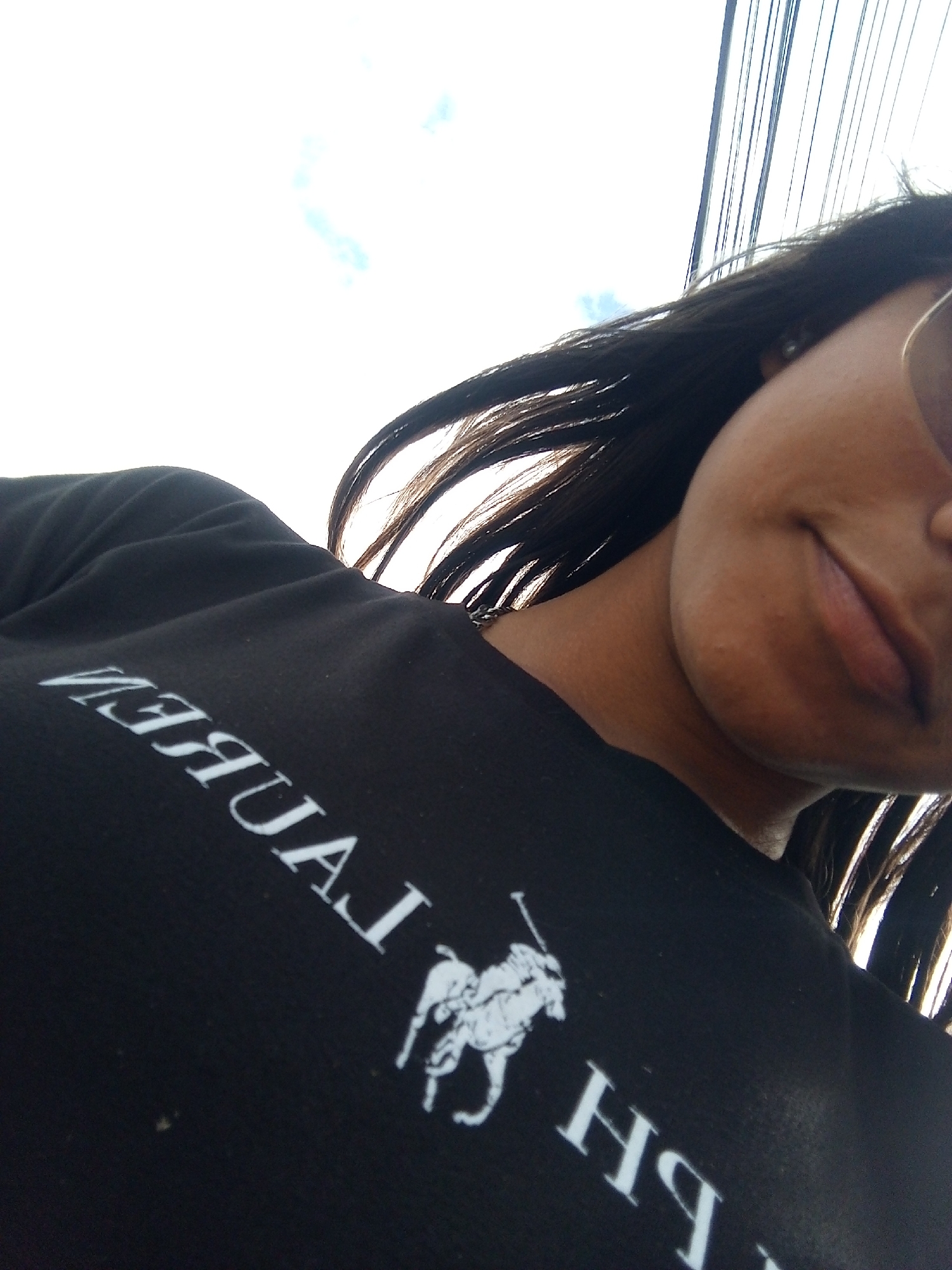 A woman with long, dark hair is pictured from a low angle. She wears sunglasses, obscuring her eyes, and a black Ralph Lauren t-shirt with the brand name printed in white. The background shows utility wires against a pale sky with a few clouds. The location is consistent with an outdoor setting, potentially in Juan Griego, Venezuela, based on the possibility of identifying the location using additional details.