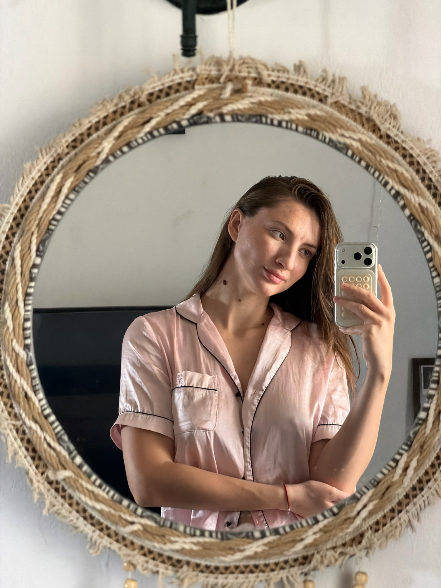 A woman is taking a selfie in a round mirror with a woven frame. She is wearing pink pajamas and has her hair down. The mirror reflects her upper body and face. She is holding a phone in her right hand. The background is a white wall and a dark object, possibly a television, is visible in the reflection. The image appears to have been taken indoors. There are no discernible elements that place the location in Sharm El Sheikh, Egypt.