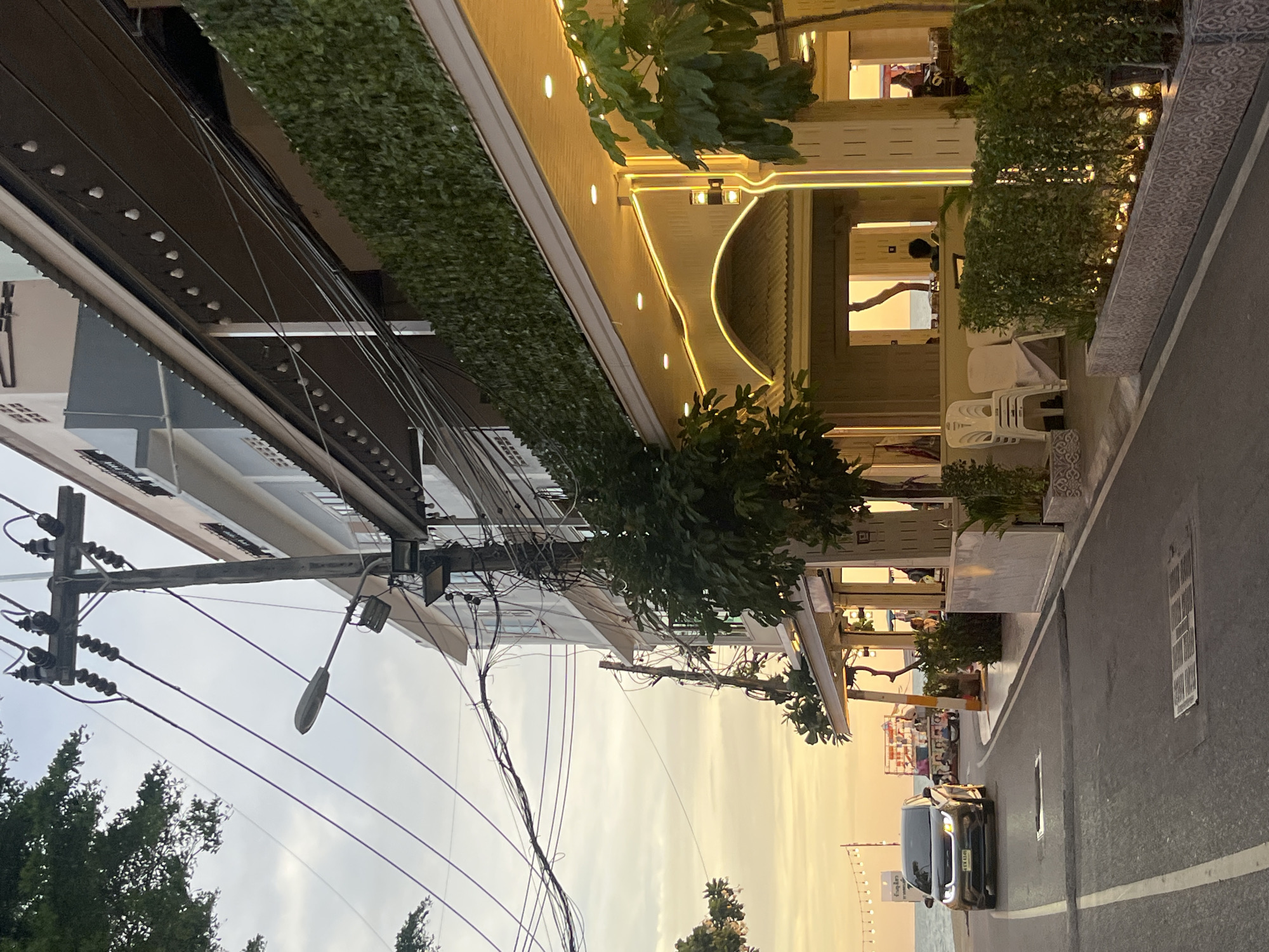 This image captures a street scene in Pattaya City, Thailand. The view is oriented such that the road runs from bottom right to the middle of the frame. A car is positioned on the right side of the road. Overhead, there are numerous electrical wires connected to a utility pole on the left side. The foreground includes trees and buildings on both sides of the road. The building on the right has decorative lighting and greenery. People are visible inside of the building on the right. The background shows a beach.