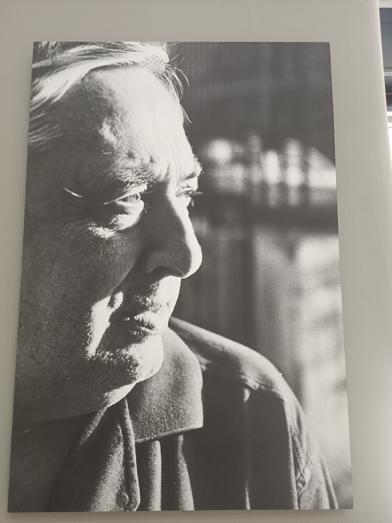 A black and white, waist-up, profile portrait of an older man is displayed on a wall. The man is looking to the right and his face is illuminated by light from the left, casting shadows on the right side of his face. He has white hair that appears windswept. He is wearing a collared shirt. The background is blurred and appears to be an indoor setting with indistinct shapes of light and dark. The photograph is hung on a plain, light-colored wall.